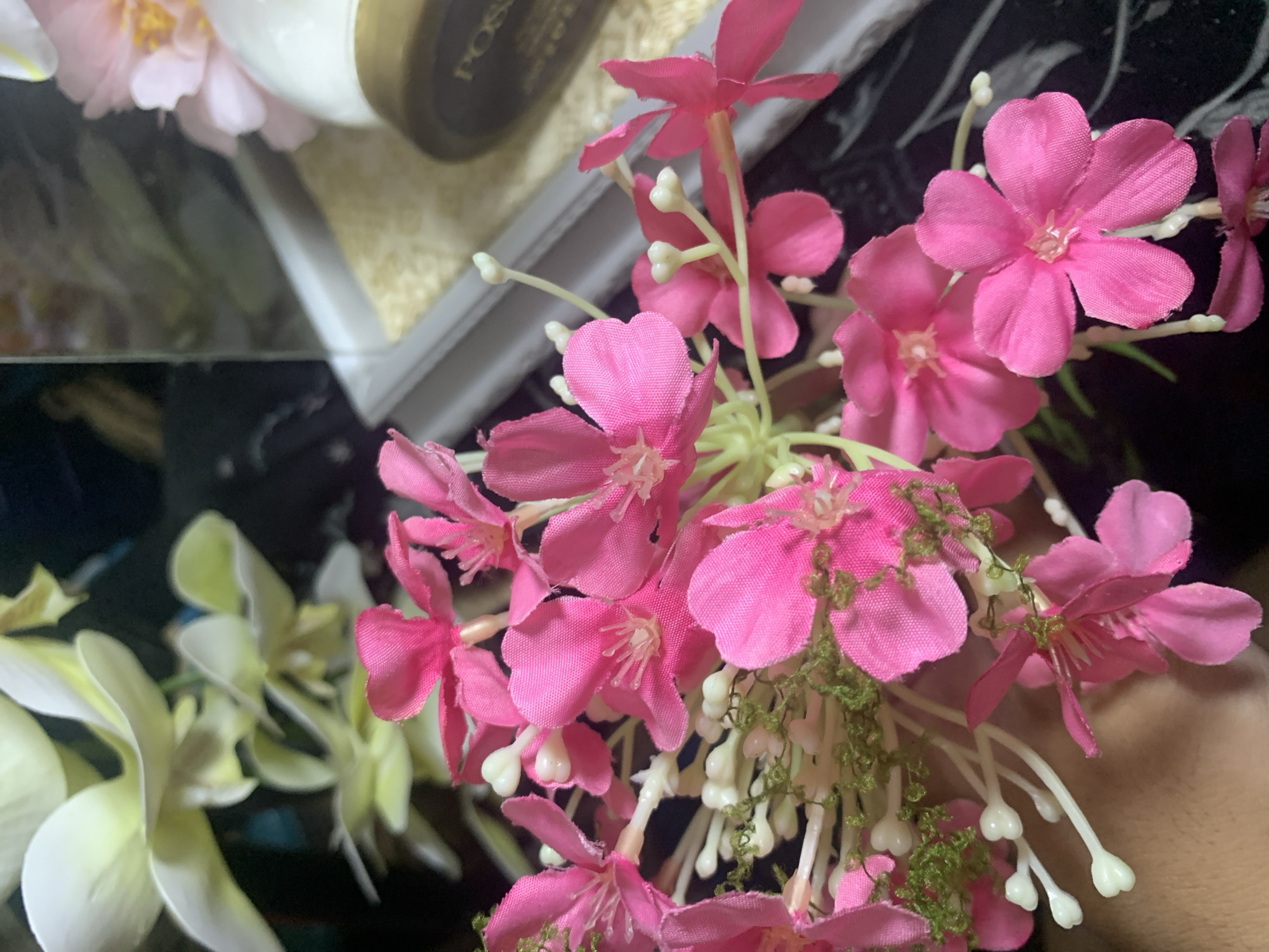 A close-up, overhead shot captures a bouquet of vibrant pink artificial flowers with delicate white bead-like accents. The flowers are intricately detailed, with soft fabric petals and thin green stems. In the background, a blurry assortment of pale yellow and white flowers adds depth. To the right, a white jar with a gold lid is partially visible, suggesting a decorative arrangement or gift set. The overall scene is bright and colorful, with a shallow depth of field that blurs the background. The image does not contain any elements that allow for the identification of a specific location in Kuala Pembuang, Indonesia, or any individuals.