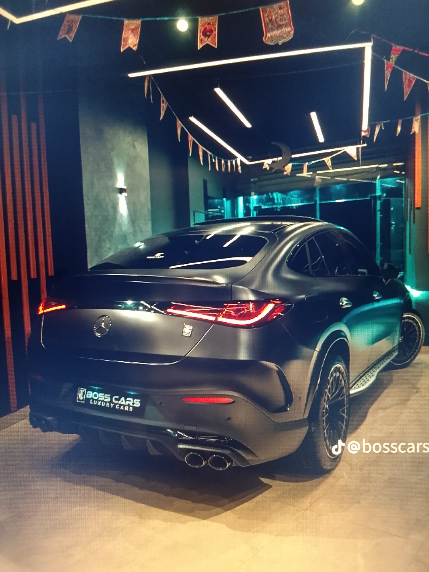 A satin black or matte dark grey Mercedes-Benz SUV coupe, likely a Mercedes-AMG GLE Coupe, is prominently displayed indoors within what appears to be a luxury car showroom or dedicated garage. The vehicle is positioned in a rear three-quarter view, angled slightly towards the right, showcasing its sleek, sloping roofline, illuminated horizontal LED taillights, quad exhaust tips, and multi-spoke black wheels. A metallic Mercedes-Benz emblem is centered on the tailgate, and to its right, a small square emblem is visible. A side step or running board extends along the passenger side. The rear license plate area features a custom plate holder displaying "BOSS CARS LUXURY CARS" with an associated logo.
The surrounding environment consists of dark walls; the left wall features vertical orange-brown slatted panels, while the wall behind the car is a textured dark grey. Overhead, a dark ceiling is fitted with bright, linear LED strip lights arranged in geometric patterns, providing primary illumination. Ornamental bunting, featuring small, rectangular patterned flags, is strung across the ceiling. A crescent moon-shaped ornament hangs from the ceiling in the mid-background. In the far background, a large glass partition or window reveals an area with distinct blue-green ambient lighting. The floor beneath the vehicle is a light-colored, possibly tiled or polished concrete surface. No individuals are present in the scene. A TikTok logo and the handle "@bosscars" are digitally overlaid in the bottom right corner of the image. The location is Unknown, Unknown.