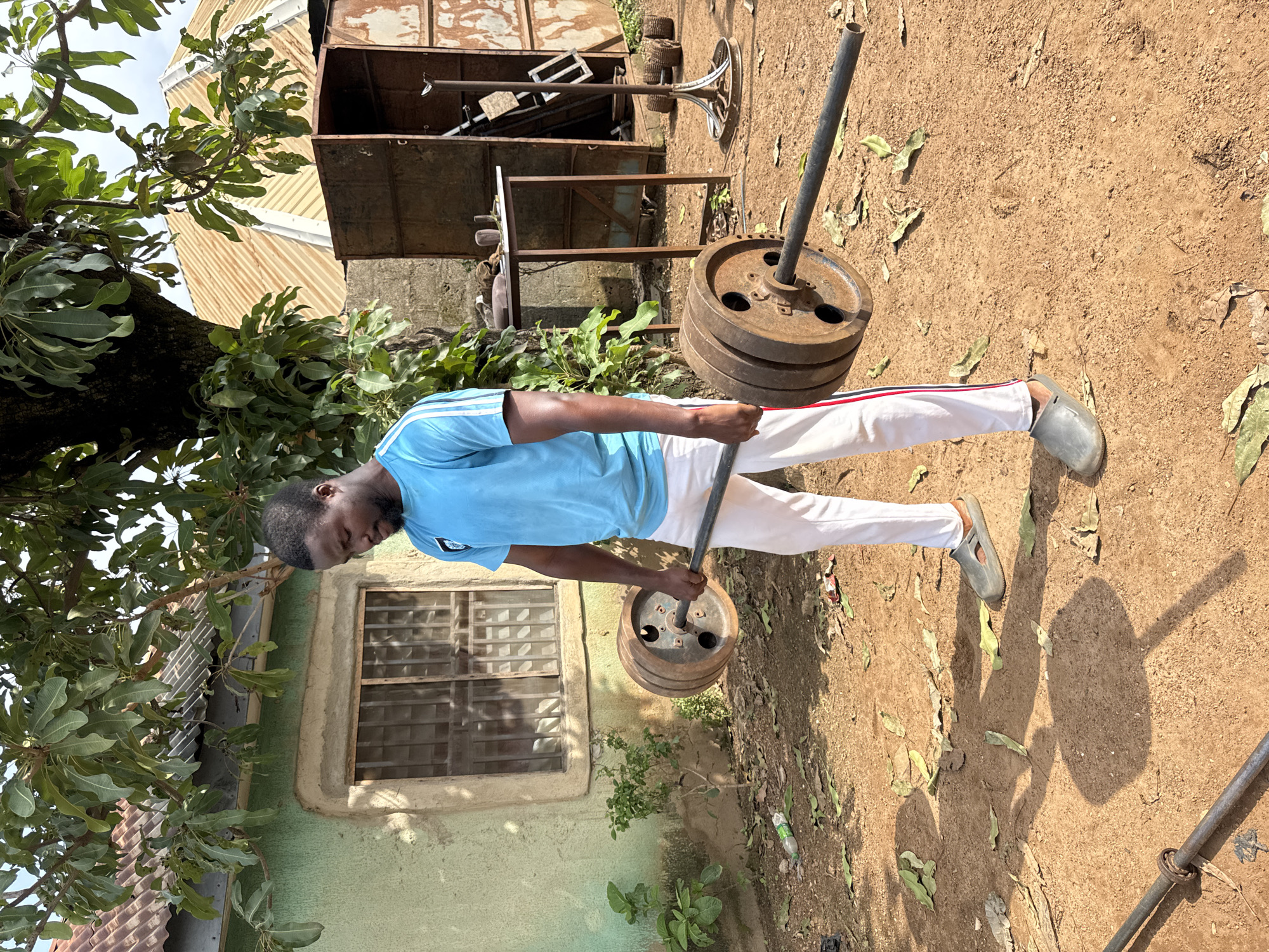 In Akwanga, Nigeria, a Black man with short dark hair and a beard is outside, lifting weights. He is wearing a bright blue t-shirt with white stripes on the sleeves and white pants with red and blue stripes down the sides. He is holding a barbell with weights on each end. The ground is dirt and scattered with dry leaves. Behind him is a light green building with a window that has metal bars. There is also a large tree and a rusty metal structure with a table in front of it.