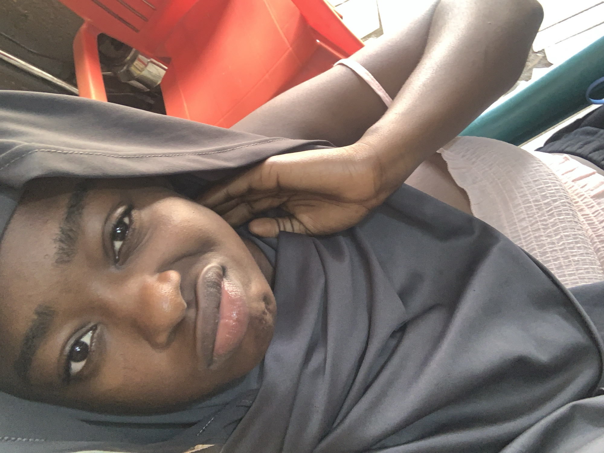 A close-up, angled photograph captures a Black woman with dark hair, dark eyes, and full lips. She is lying down, draped in a grey garment that partially covers her head. Her right hand is near her face. In the background, a red plastic chair and a cylindrical green object are visible. A white, patterned fabric is also present. The location is identified as Rafin Par, Nigeria.