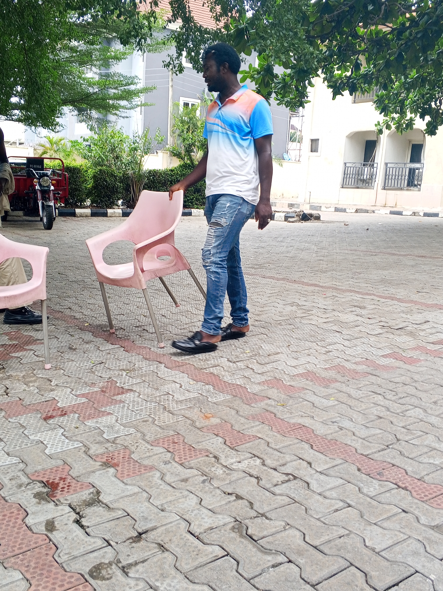 A man stands next to a pink plastic chair on a paved surface in Lokoja, Nigeria. The paving consists of interlocking, dog-bone-shaped concrete blocks in alternating rows of gray and red. The man is Black, wearing a white t-shirt with blue and peach accents, ripped blue jeans, and black loafers. He is looking to his right and his left hand rests on the back of the chair. Another chair is visible to the left, occupied by a person wearing black shoes. Behind them, a red three-wheeled vehicle, possibly a commercial tricycle, is parked near a curb. The background includes a mix of buildings, trees, and shrubs. The buildings appear to be multi-story residential or commercial structures, some with balconies. The scene is outdoors during daylight.