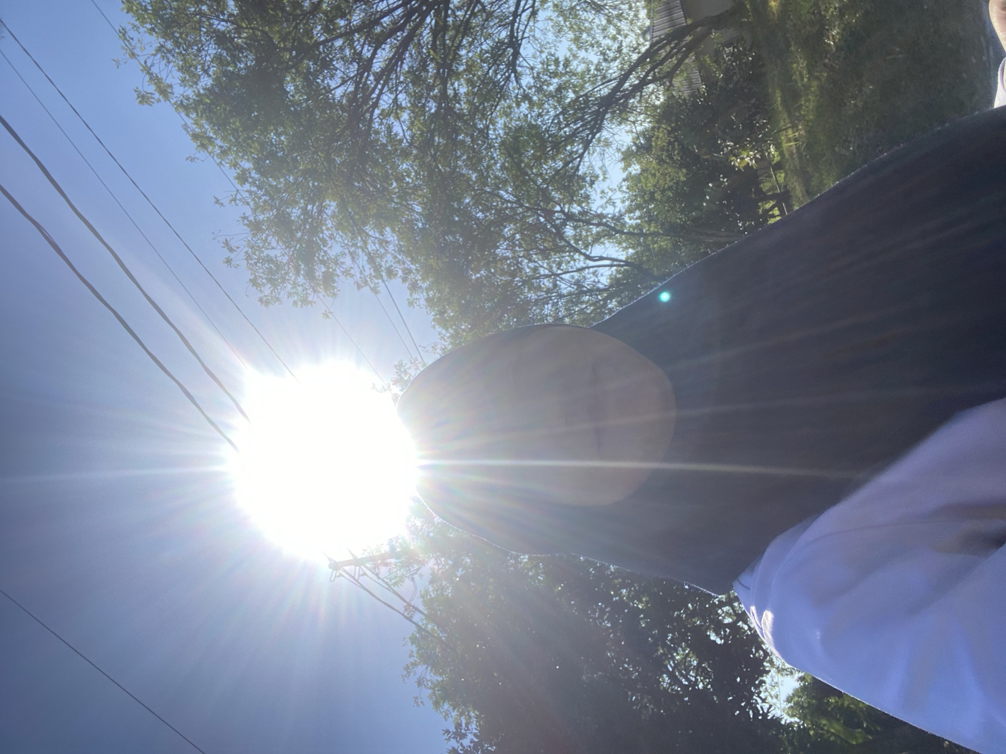 A selfie is taken outdoors in Banjarbaru, Indonesia, on a sunny day, with the sun directly overhead and bright rays emanating from it. The subject, wearing a black hijab and a white top, is partially obscured by the sun's glare. In the background, lush green trees and power lines are visible under a clear blue sky. The overall impression is one of a warm, bright day in a natural, possibly suburban or rural setting within the city.