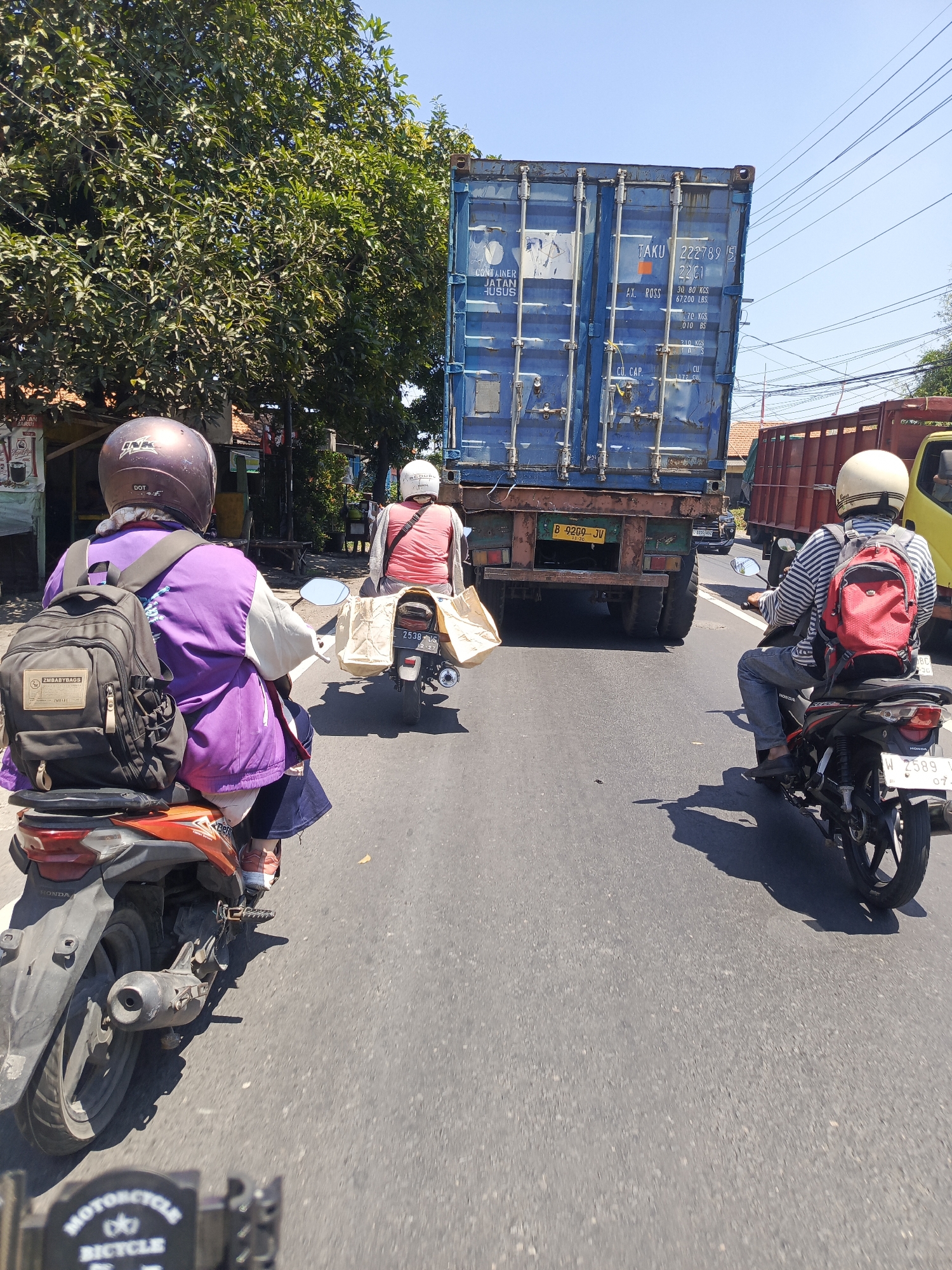 This image depicts a street scene in Ngelom, Indonesia, with a focus on traffic. In the foreground, two individuals are riding motorcycles. The motorcycle on the left is ridden by a person wearing a purple jacket and a dark helmet, carrying a black backpack. The motorcycle on the right is ridden by a person wearing a striped shirt, a white helmet, and a red backpack. Both motorcycles are in motion on an asphalt road, with one carrying a large shopping bag on its rear. In the background, a large blue shipping container truck is prominently featured, dominating the center of the frame. The truck has various markings and labels on its side. To the right of the container truck, a red truck is partially visible. The environment is characterized by a clear blue sky and lush green trees lining the sides of the road. The overall scene suggests a typical day of road traffic in an urban or semi-urban Indonesian setting.
