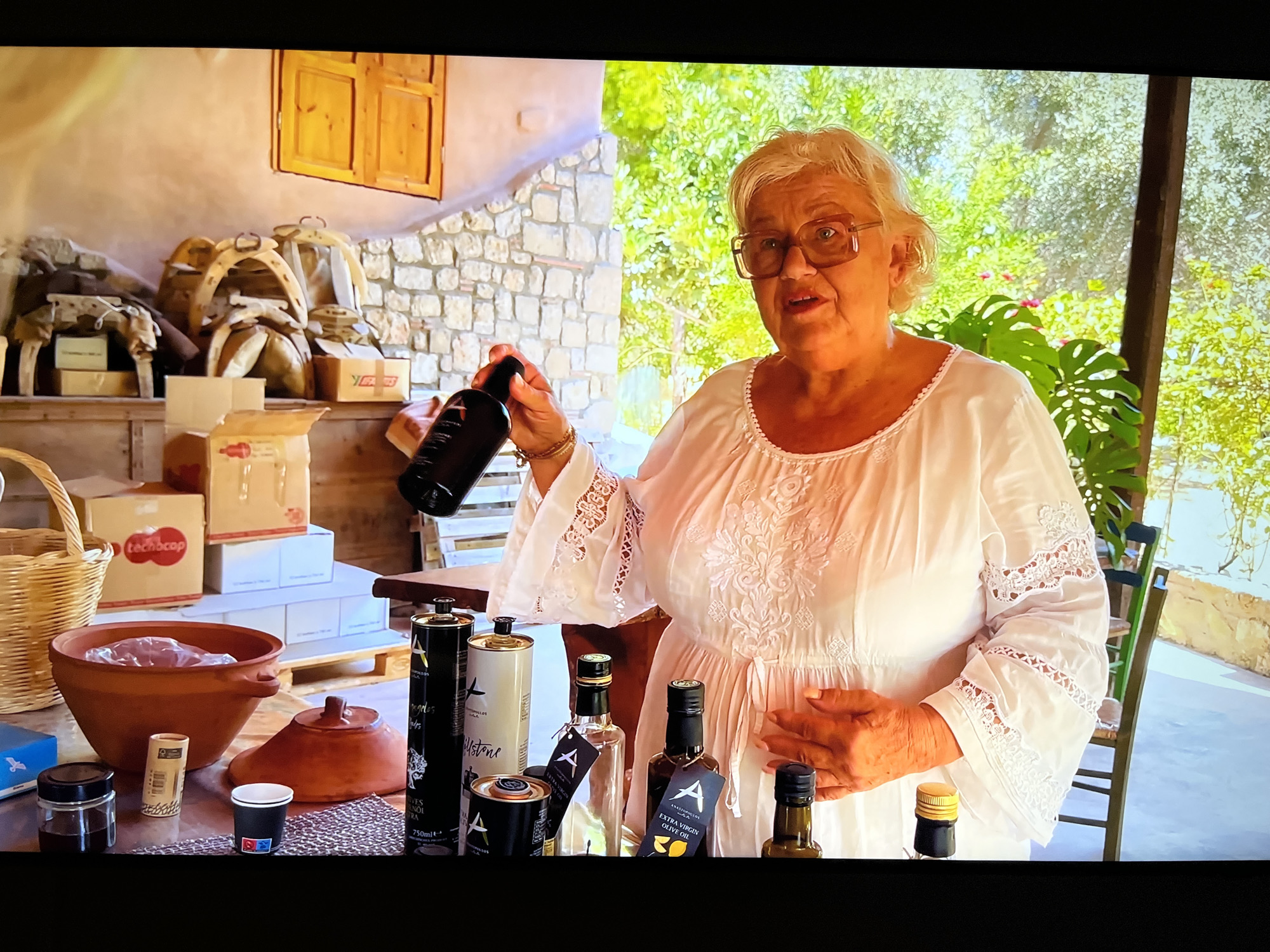 A middle-aged woman with short blonde hair and glasses, wearing a white embroidered dress, stands in what appears to be an outdoor setting, possibly a kitchen or a covered patio, with lush green foliage visible in the background. She is holding a dark bottle in her right hand and gesturing with it. In front of her on a table, there are several bottles, primarily of olive oil, and a terracotta bowl containing food. The background shows a stone wall and wooden elements, suggesting a rustic or traditional environment. There are no visible indications of the city of Kharkiv, Ukraine, in the image.