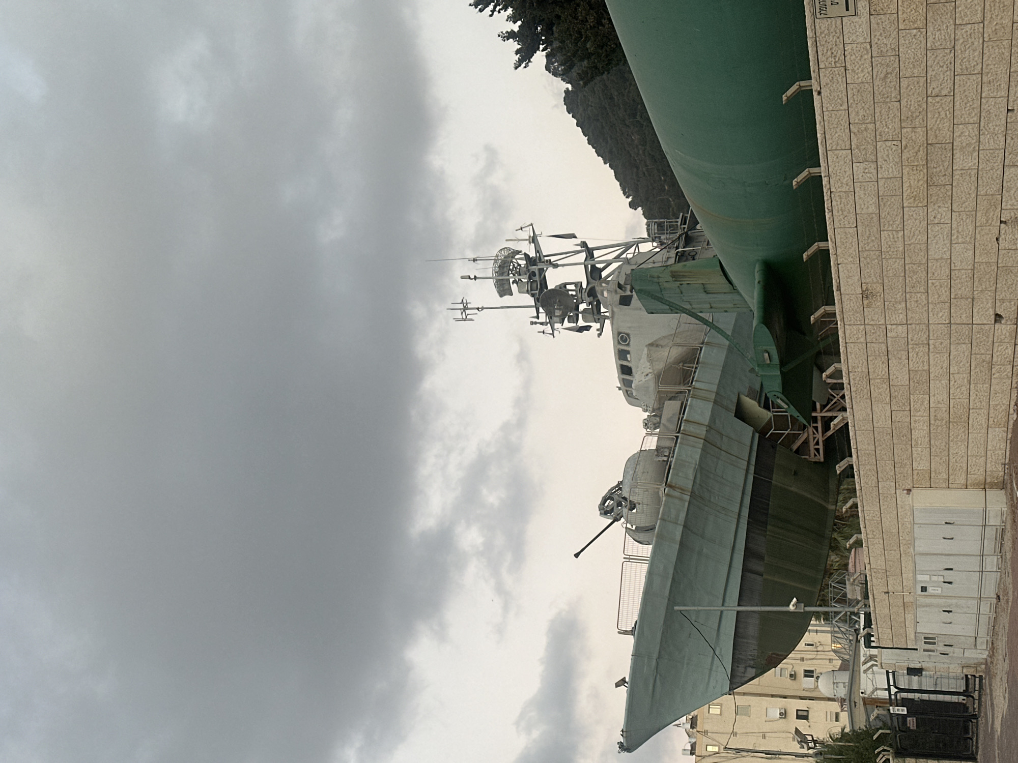 The image captures a naval vessel, a former missile boat, prominently displayed as an exhibit. The boat, painted in a muted green, is elevated and positioned behind a light-colored brick wall. Its sleek hull and superstructure, equipped with various antennas and a gun turret, are visible against a cloudy sky. To the right of the boat, a large, cylindrical green object, possibly a torpedo or a submarine hull, is partially visible. In the background, a dense green hillside dotted with trees rises behind the exhibit. To the left, a modern apartment building with multiple windows stands, suggesting an urban setting. The foreground shows a metal fence and electrical boxes near the brick wall. The location is identified as the National Maritime Museum in Haifa, Israel, where the INS Hanit, a Sa'ar 4-class missile boat, is displayed. There are no people visible in the frame.