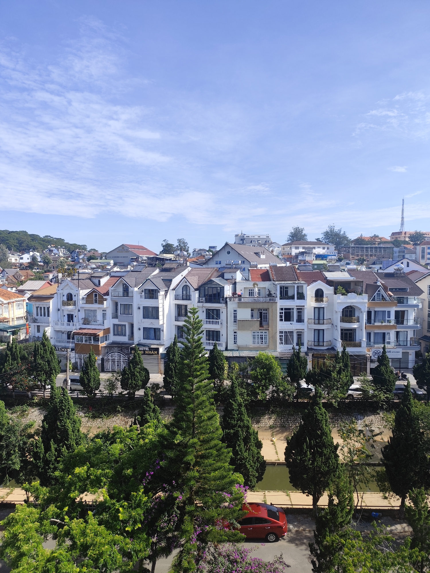 The image depicts a street scene in Da Lat, Vietnam, characterized by its distinctive architecture and lush greenery. In the foreground, an abundance of trees and shrubs, including several coniferous evergreens, partially obscures the view of a small river or canal. A red sedan is parked along the adjacent street.
Behind the trees, a row of closely-spaced townhouses or villas stands. These buildings exhibit a mix of architectural styles, with white facades, tiled roofs, and balconies. Some of the buildings have signage, indicating they are commercial properties or hotels.
In the background, a hillside is covered with a dense collection of buildings, blending residential and commercial structures. A tall communications tower rises above the cityscape in the distance. The sky is predominantly blue, with scattered clouds. The overall scene suggests a tranquil, scenic urban setting.