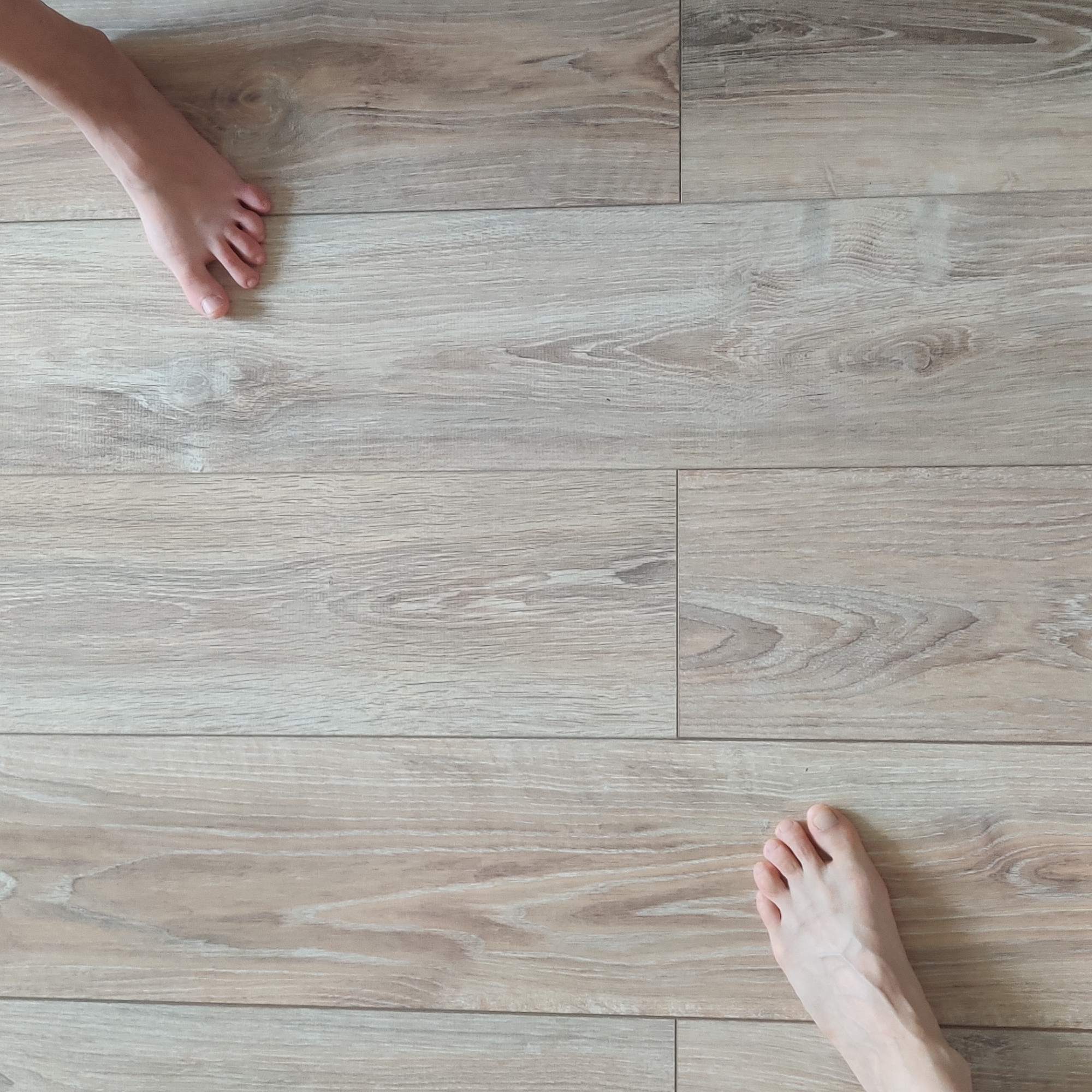 The image shows two bare feet on a light-colored wooden floor. The feet are positioned diagonally opposite each other in the frame. The floor has a visible wood grain pattern with alternating light and dark tones. The lighting suggests an indoor environment. There is no discernible connection to Yekaterinburg, Russia, as the image provides no contextual clues to location.