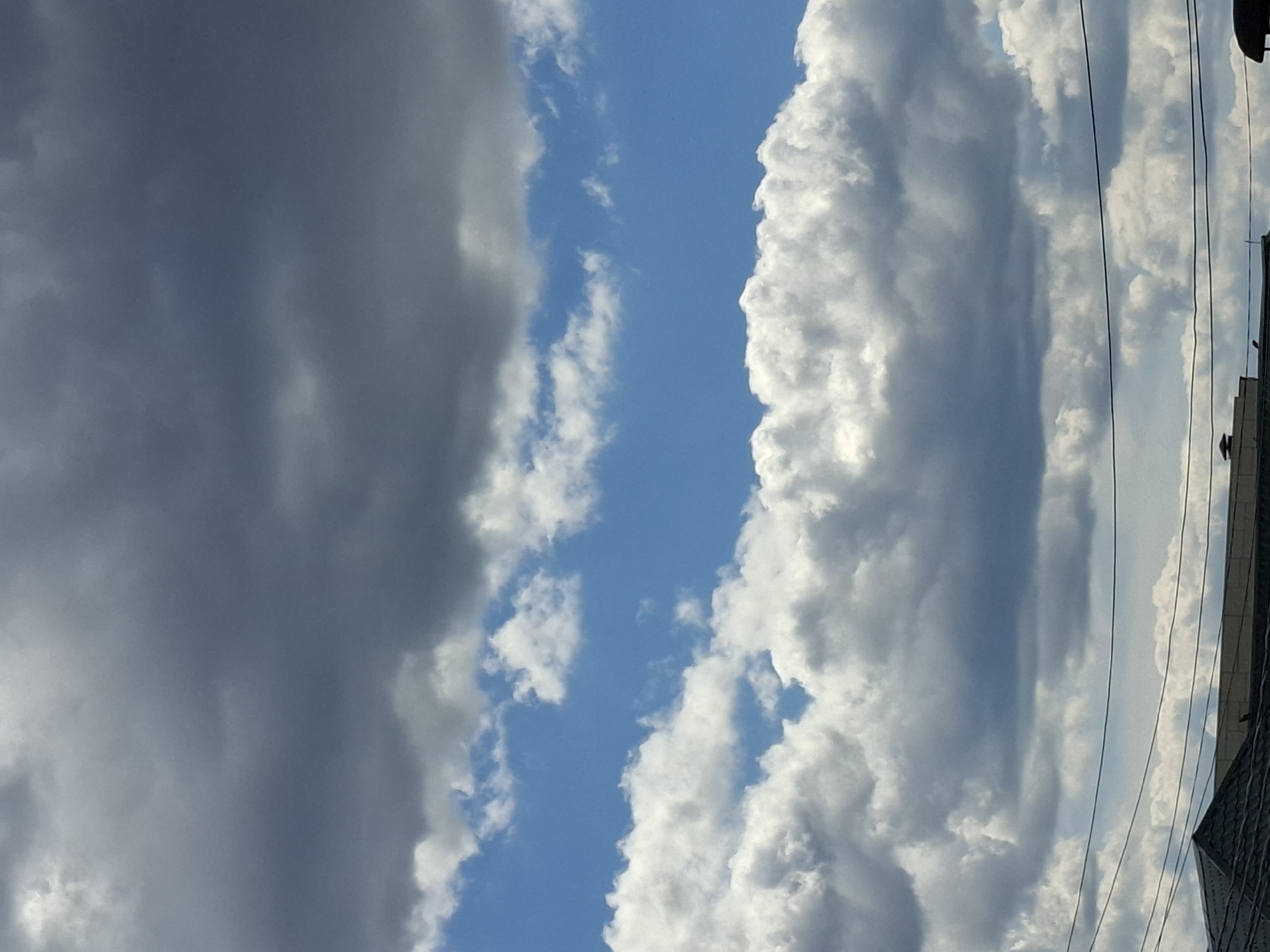 The image shows a vertical view of the sky filled with dramatic clouds. On the left, dark, heavy storm clouds dominate, obscuring much of the blue sky. To the right of the storm clouds, a bright strip of clear blue sky is visible, interspersed with scattered white, fluffy clouds. Further to the right, massive, white, and light gray cumulus clouds are prominent, catching the sunlight and creating a sense of depth and texture. These clouds appear bright and voluminous.
In the far right portion of the image, the edge of a building is visible, along with several dark, thin electrical wires that stretch diagonally upwards, suggesting an urban or semi-urban environment. The roofline of the building is also visible, appearing dark and angular.
The contrast between the dark storm clouds and the bright, sunlit clouds creates a dynamic and somewhat ominous atmosphere. The presence of the building and wires indicates this is likely a daytime scene, possibly late afternoon or early evening, given the quality of light and the contrast in cloud formations. The weather appears changeable, with a storm brewing on one side and clear skies elsewhere. There is no visible text in the image.