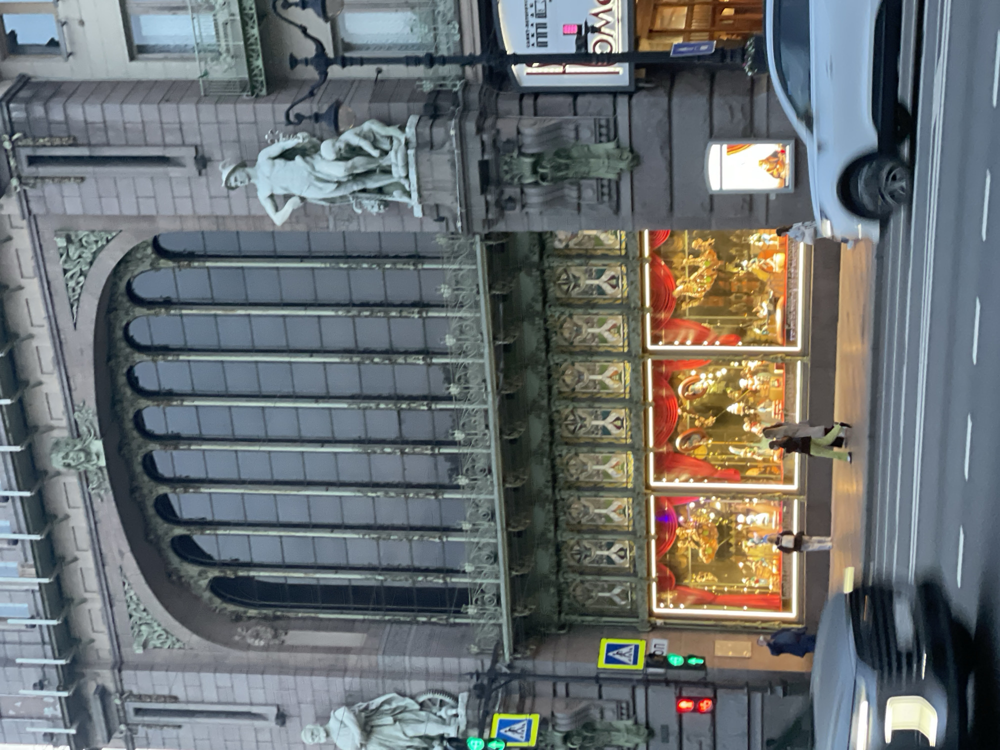 The image depicts a section of Nevsky Prospect in Saint Petersburg, Russia, specifically a portion of the Eliseev Emporium building. The architectural features of the building are visible, including large arched windows, decorative moldings, and sculptures. Several illuminated display windows showcasing decorative merchandise can also be seen. Pedestrians are present on the sidewalk, and vehicles, including cars, are on the road. Traffic lights and crosswalk signs are also visible, indicating an active street scene.