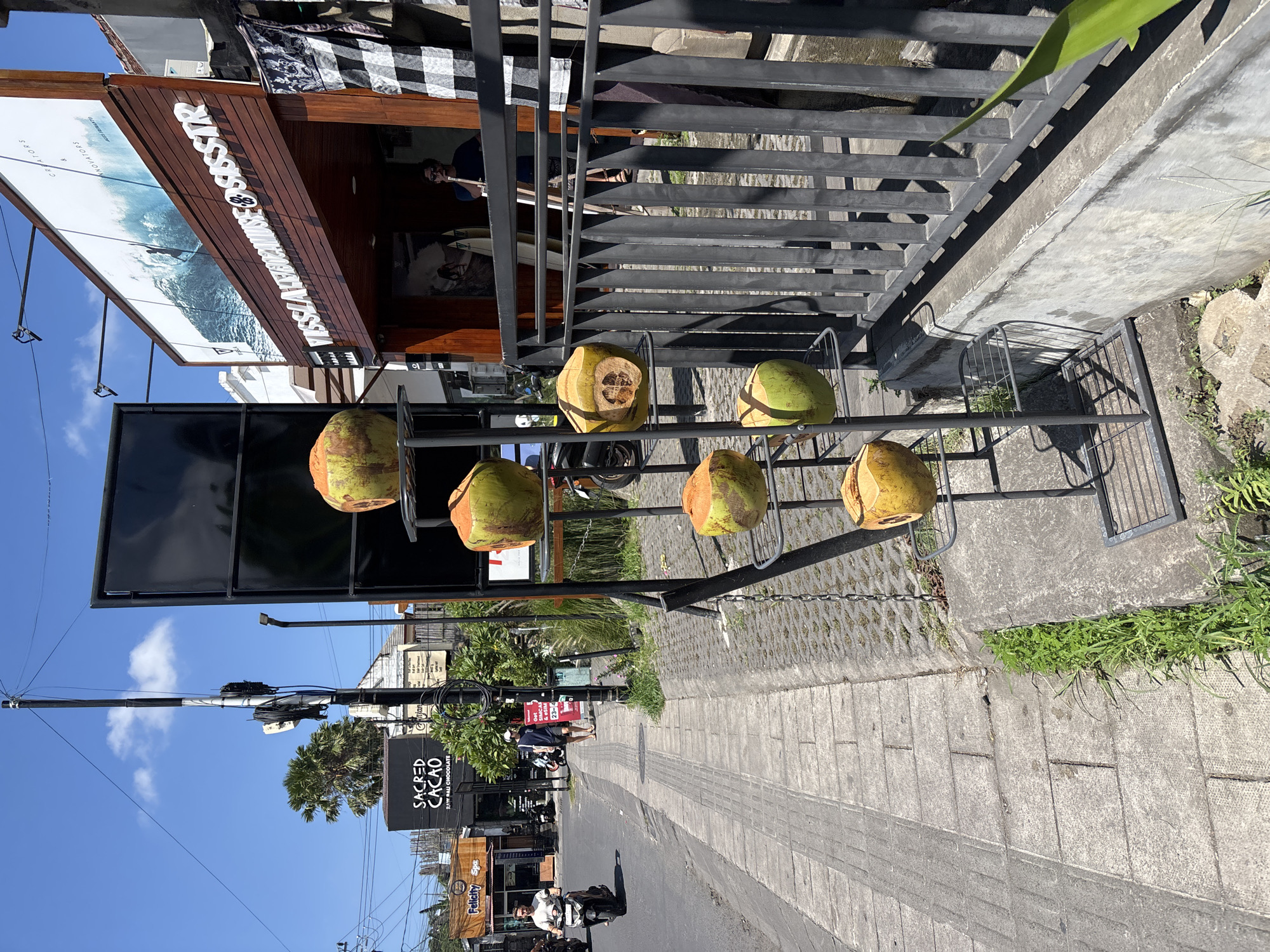 Here is a description of the image:
The image shows a street scene in what appears to be North Kuta, Indonesia. A metal rack displays six coconuts, some cut open. Behind the rack is a building with a black and white awning. To the left, signage indicates businesses like "Sacred Cacao" and "Felicity Spa" on the street. People are seen riding scooters and walking on the street. The sky is blue and clear. Buildings, trees, and power lines line the street. "Vissla" is printed on the side of one of the businesses.