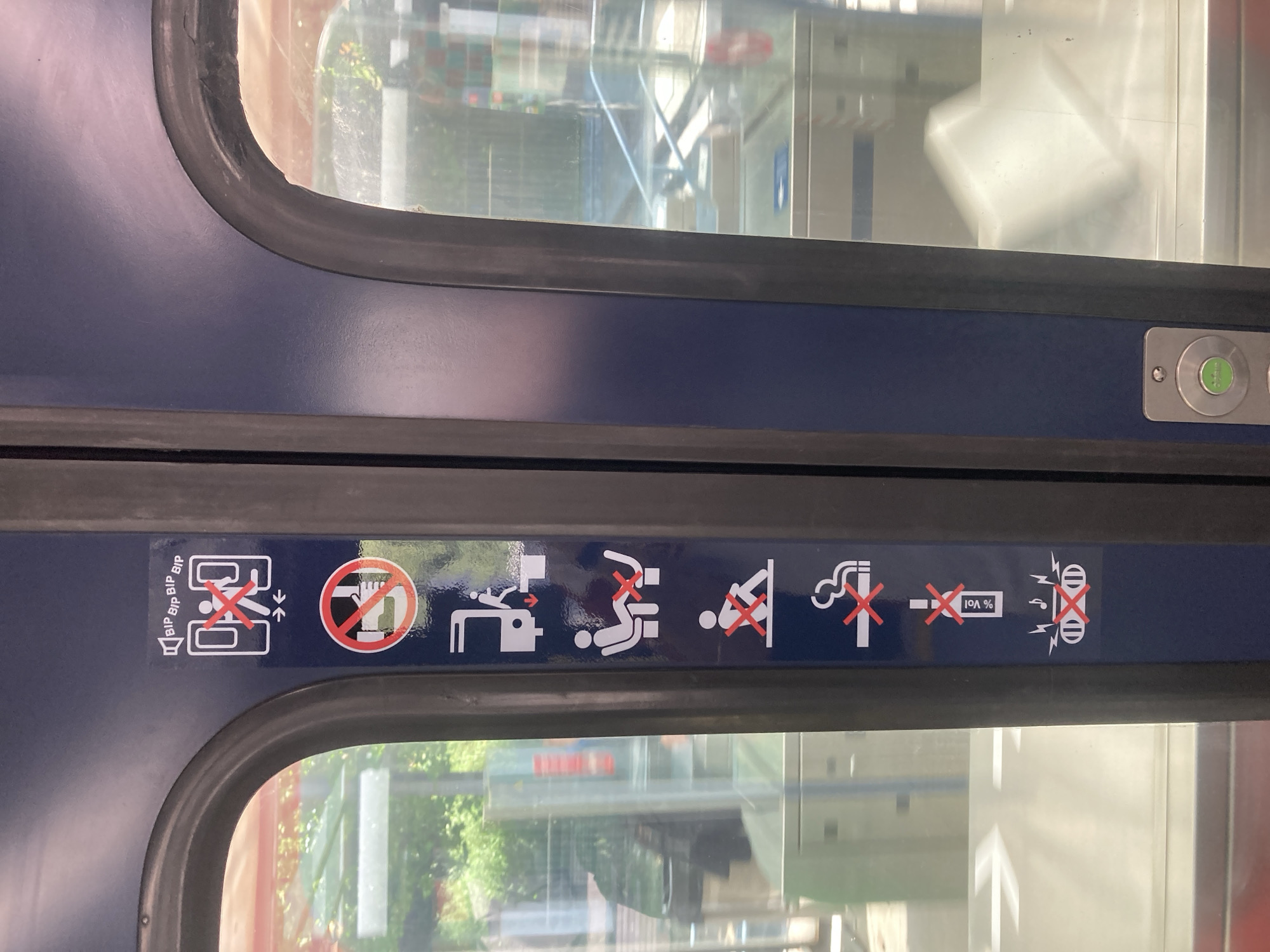 The image displays a section of a dark blue door, likely on a public transport vehicle like a train or metro, featuring two windows with black rubber seals. A series of white pictograms with red crosses indicating prohibitions are displayed horizontally on the blue panel between the two windows.
From left to right, the pictograms convey the following messages:
1. **"BIP BIP BIP BIP" text with a speaker icon and a crossed-out person trapped between closing doors:** This warns against obstructing the closing doors.
2. **A red circle with a diagonal line over an open hand:** This signifies "do not touch" or "keep hands clear."
3. **A crossed-out person next to a shopping cart or stroller with a downward arrow:** This indicates that the area, possibly the door or aisle, should not be blocked by luggage or strollers.
4. **A crossed-out person squatting or sitting on the floor:** This prohibits sitting on the floor or in non-designated seating areas.
5. **A crossed-out person with their feet elevated against a surface:** This prohibits placing feet on seats, walls, or windows.
6. **A crossed-out smoking cigarette:** This indicates "no smoking."
7. **A crossed-out bottle with "% Vol":** This prohibits the consumption of alcoholic beverages.
8. **A crossed-out person wearing headphones with lightning bolts emanating from them:** This likely means "no loud music" or "no use of electronic devices at high volume."
To the right of the pictograms, a silver panel is visible, containing a circular green button, possibly for opening the door or an emergency function, next to another similar button that is not fully visible.
The windows reflect outdoor scenery, including parts of buildings, trees, and what appears to be another section of the station or vehicle interior, suggesting it is daytime. An arrow pointing upwards is also faintly visible in a reflection. The overall setting is an interior of a public transport vehicle, conveying a set of rules and warnings to passengers.