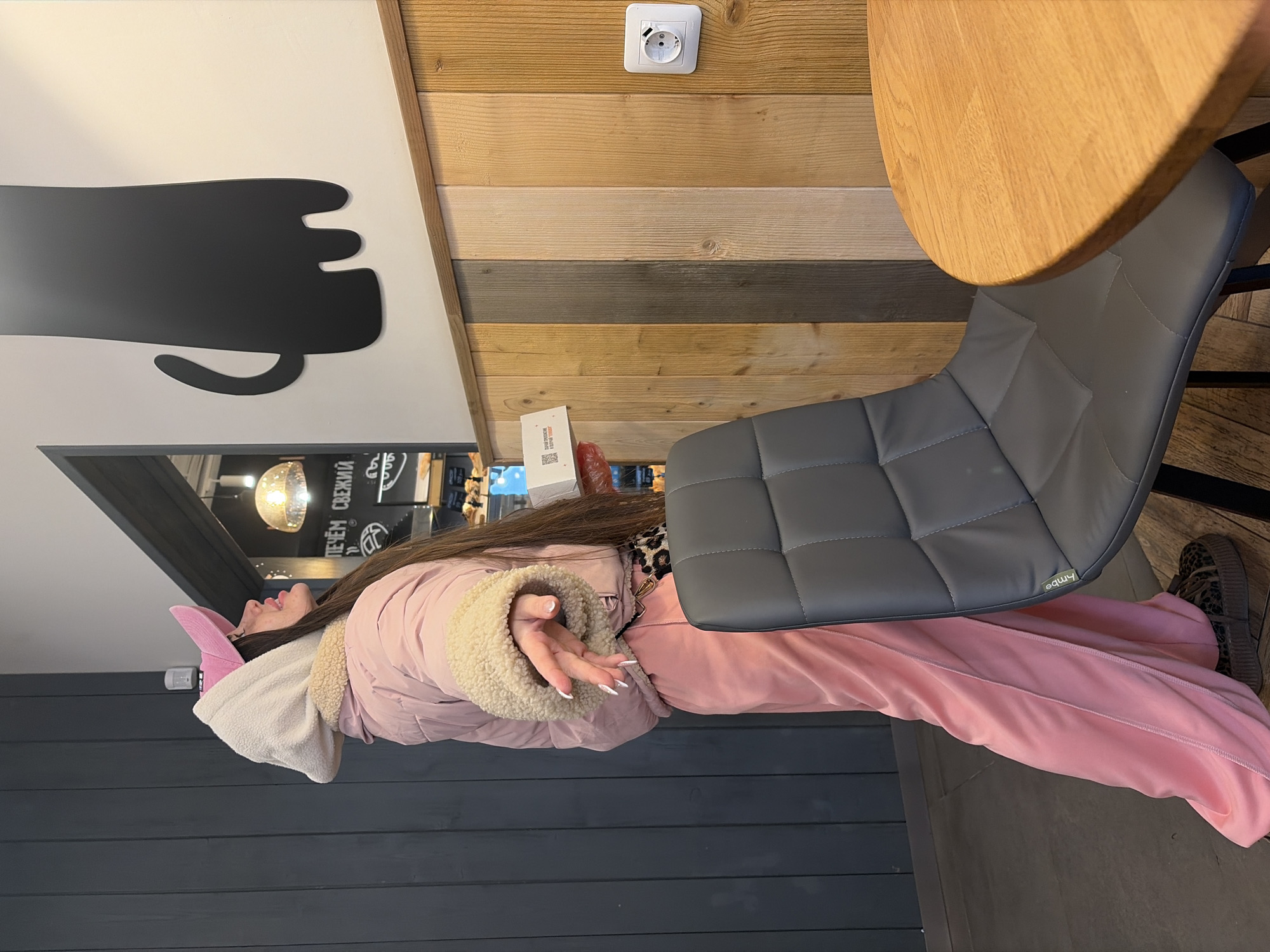 A woman wearing a pink jacket, pants, and shoes is sitting on a gray chair. The woman is also wearing a white hoodie with pink bunny ears attached. She is sitting at a round wooden table. On the wall, there is a graphic of a black mug with a handle. The wall is decorated with alternating wood paneling, and there is a white electrical outlet on the wall. The location is in the city of Saint Petersburg, Russia.