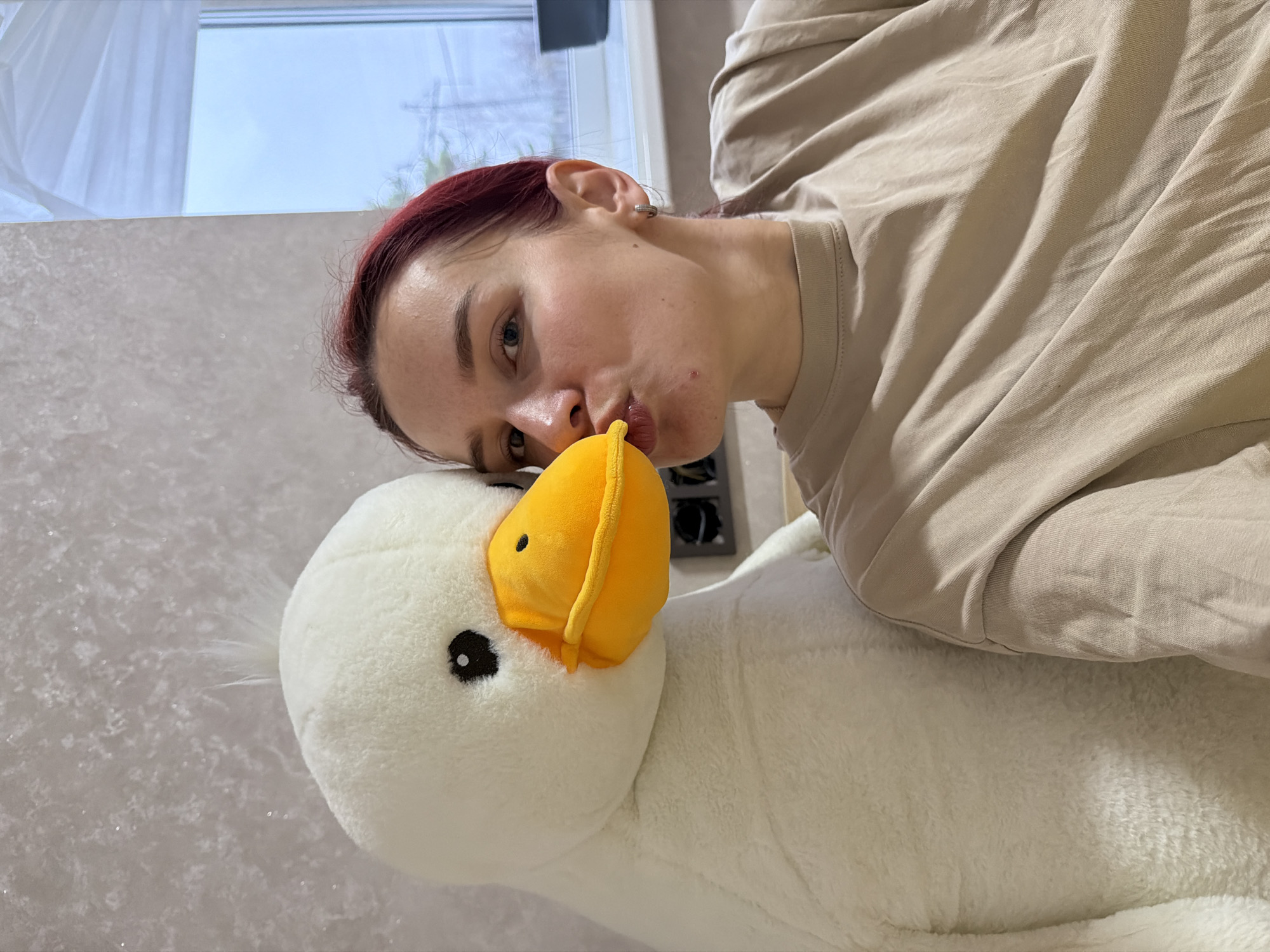 A young woman with red hair is positioned sideways, appearing to kiss the beak of a large, plush white goose with a yellow beak. She is wearing a beige t-shirt and a silver hoop earring. The background includes a window with a view of an overcast sky, and what appears to be a wall with a textured or patterned surface. The setting seems to be indoors. The city of Chaikovsky, Russia is not discernible from the given visual information.