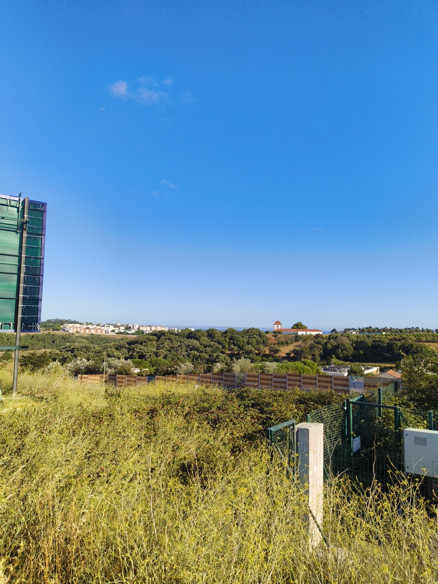 The image shows a wide-angle outdoor view of a landscape on a clear, sunny day. In the foreground, there is tall, dry, yellow-green grass and some bushes. A concrete post stands to the right of the frame, partially obscured by the vegetation. Behind the grass, there is a green metal fence with a gate. A gray electrical box is mounted on a fence post to the right. Further back, there is a row of trees and a wooden fence or barrier. Beyond the trees, a town or city is visible with many multi-story buildings. In the distance, a body of water, likely the ocean, can be seen. On a hill in the middle distance, there is a building with a red-tiled roof and a white tower, possibly a church or historical structure. To the far left of the image, a large, modern, green-paneled structure is partially visible, suggesting the presence of an industrial or commercial facility. The sky is a clear, vibrant blue with a few small, wispy clouds and a distant airplane. The overall scene depicts a semi-urbanized area with abundant vegetation and a view towards the coast. The location is identified as Cascais, Portugal.