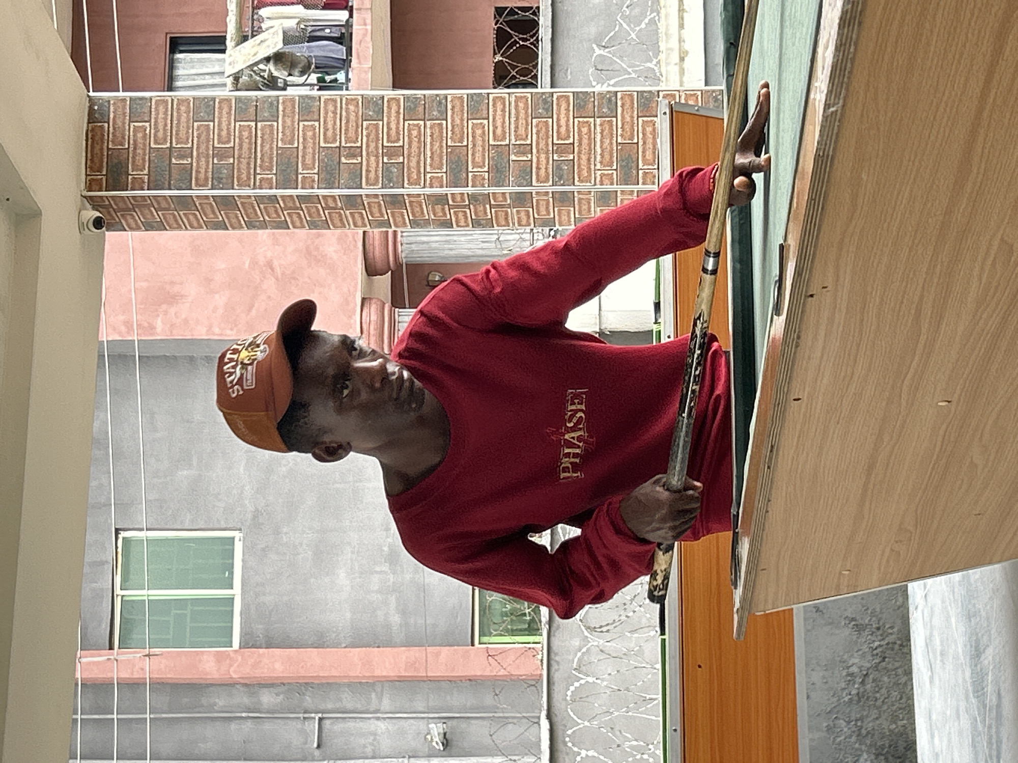 A young Black man wearing a maroon long-sleeved shirt and an orange baseball cap stands behind a green pool table, his right hand holding a cue stick poised to strike a ball. The man is in the foreground, his gaze focused on the table. In the background, a grey building with a pink vertical beam is visible, along with a small green-paned window. A brick-patterned pillar stands to the right of the man. Further back, another building with pink walls and a brown-tiled roof is present. Fencing with barbed wire is positioned in front of the grey building. The environment appears to be an outdoor covered area, possibly a residential courtyard or a public space in Ihiagwa, Nigeria.