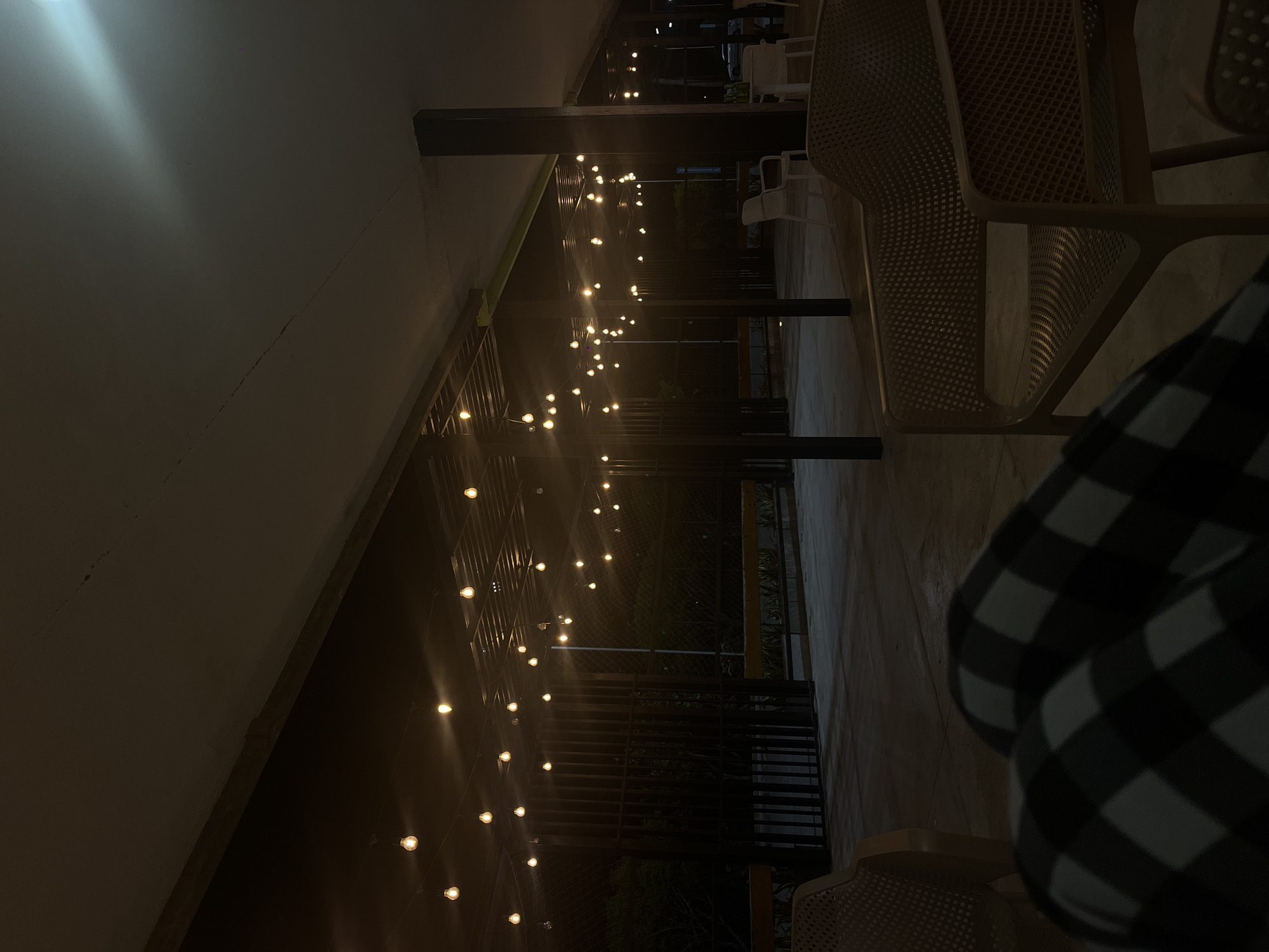 This nighttime image depicts a semi-outdoor or covered patio area, warmly illuminated by numerous string lights. In the foreground on the right, a person is partially visible, seated and wearing a black and white plaid top. Adjacent to them and extending into the midground are several light-colored, mesh-backed chairs, suggesting a casual seating arrangement.
The space is defined by dark support beams or columns, and overhead, strands of glowing, globe-style string lights stretch horizontally, providing the main source of light and creating a cozy atmosphere. The light-colored ceiling or canopy above shows a faint, diagonal crack or seam, with a brighter, localized light source reflecting in the upper left portion. Beyond the immediate seating area, the background is dark, indicating an outdoor environment, possibly featuring a railing or balustrade. The overall mood is calm and inviting. No legible text is visible in the image.