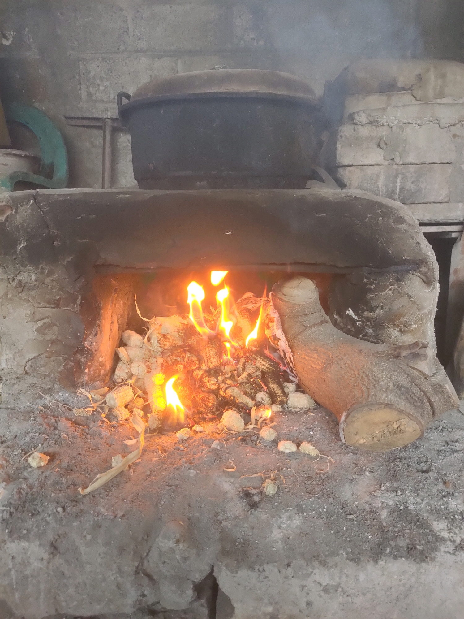 The image depicts a close-up view of a traditional wood-fired oven, likely located in Plemahan, Indonesia. The oven is constructed from rough, grey concrete and appears weathered. A large, dark pot or cauldron sits on top of the oven, partially obscuring the view of the interior. Inside the oven, a fire burns with bright orange flames, illuminating pieces of wood and ash. To the right of the fire, a thick, rough-barked log is positioned, partially entering the oven's opening, with its end cut flat, revealing the wood's grain. The surrounding ground is covered in ash and debris, consistent with an active fire. Smoke is visible rising from the top of the oven. The background shows a textured concrete wall.