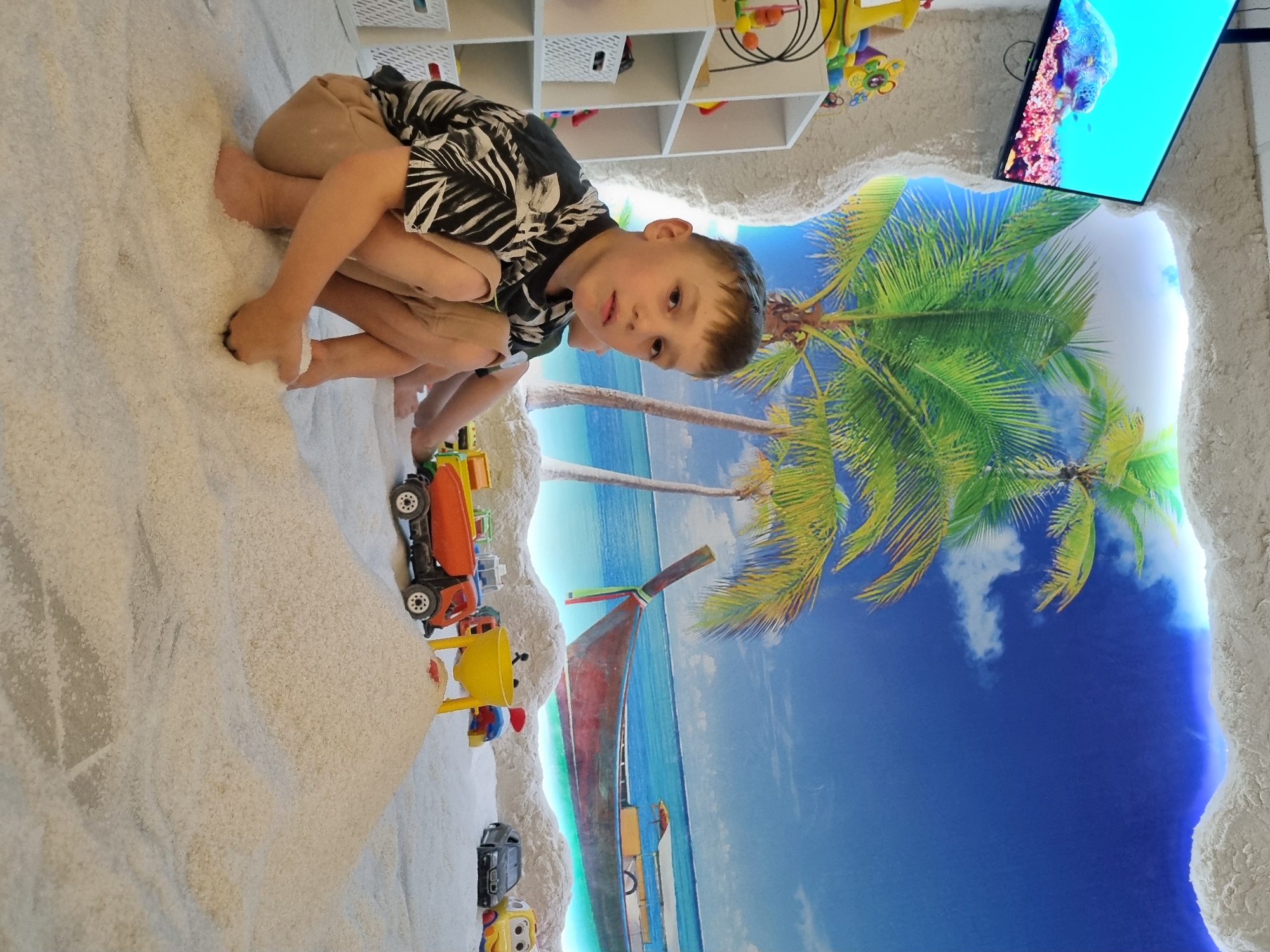 A young Caucasian boy with short brown hair and light skin is crouching in a room with a beach-themed mural and a salt-covered floor. He is wearing a black and white patterned short-sleeved shirt and tan shorts. His left leg is bent, and his right leg is tucked beneath him. His hands are in the salt, and he appears to be playing.
The room's mural depicts a tropical beach scene with palm trees, a blue sky with clouds, and a long, narrow boat on the ocean. A television screen mounted on the wall displays an image of a sea turtle. Shelving units are visible to the left of the boy, with various toys and baskets on them. In the foreground, several toy vehicles, including a red and orange dump truck, a yellow toy, and a black car, are scattered on the salt-covered floor, along with other plastic toys. The overall impression is of a children's play area, possibly a salt therapy room, given the salt-covered floor. The specific location in Chaikovsky, Russia, is not identifiable from the image alone.