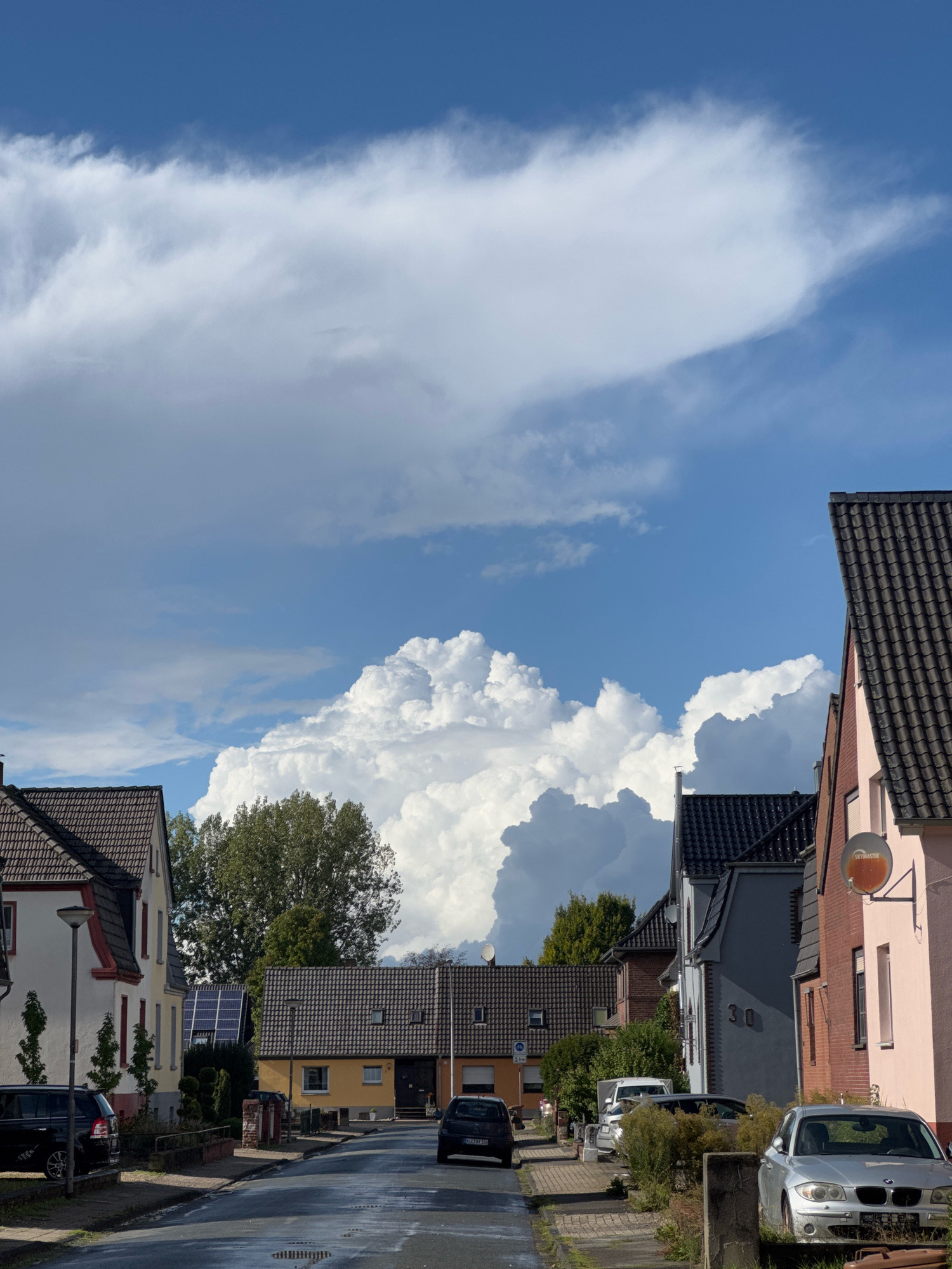 The image depicts a street in Cleves, Germany, with a row of residential houses on either side. The sky is a prominent feature, filled with dramatic, large cumulus clouds, some of which appear dark and potentially indicative of approaching rain. The street is paved and appears wet, suggesting recent rainfall. Several cars are parked along the street and on the sidewalk. Trees and greenery are visible between the houses and along the street. The architecture of the houses is varied, with different roof styles and colors. On the right side of the street, one house has a satellite dish mounted on its exterior. The overall scene conveys a somewhat overcast or post-rain atmosphere.