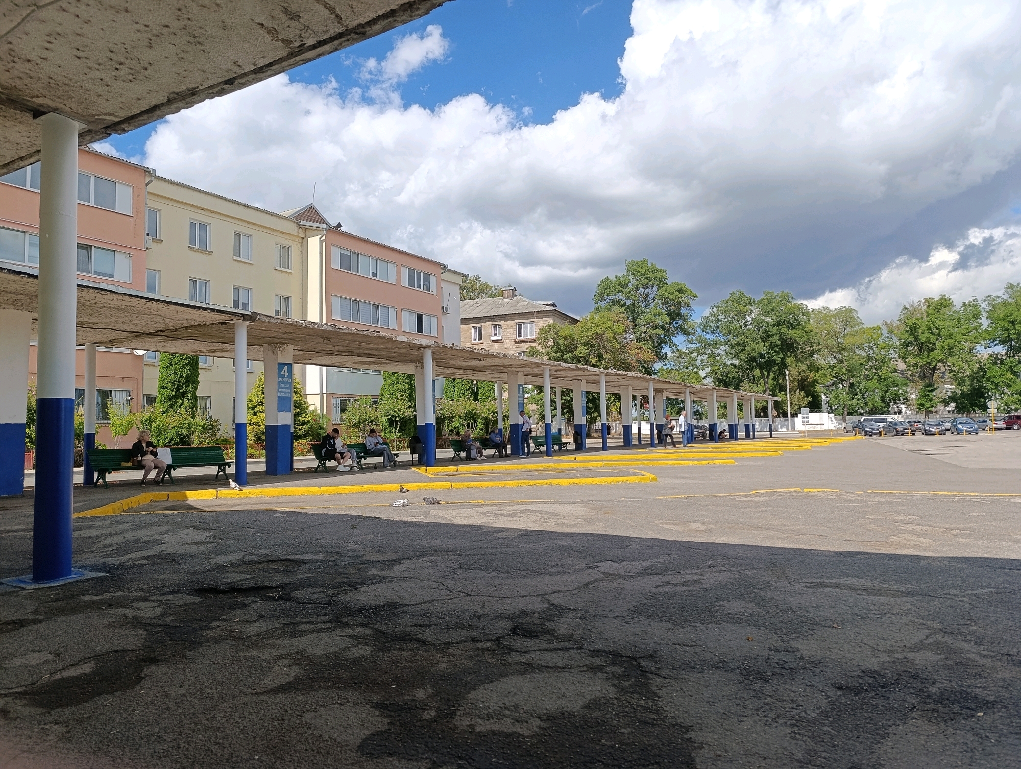 A bus stop in Bender, Moldova, features a long, covered waiting area with blue and white striped pillars. Green benches are positioned along the wall of a multi-story building, with a few people seated. The ground is paved with asphalt, marked with yellow lines. Trees and other buildings are visible in the background, under a partly cloudy sky.