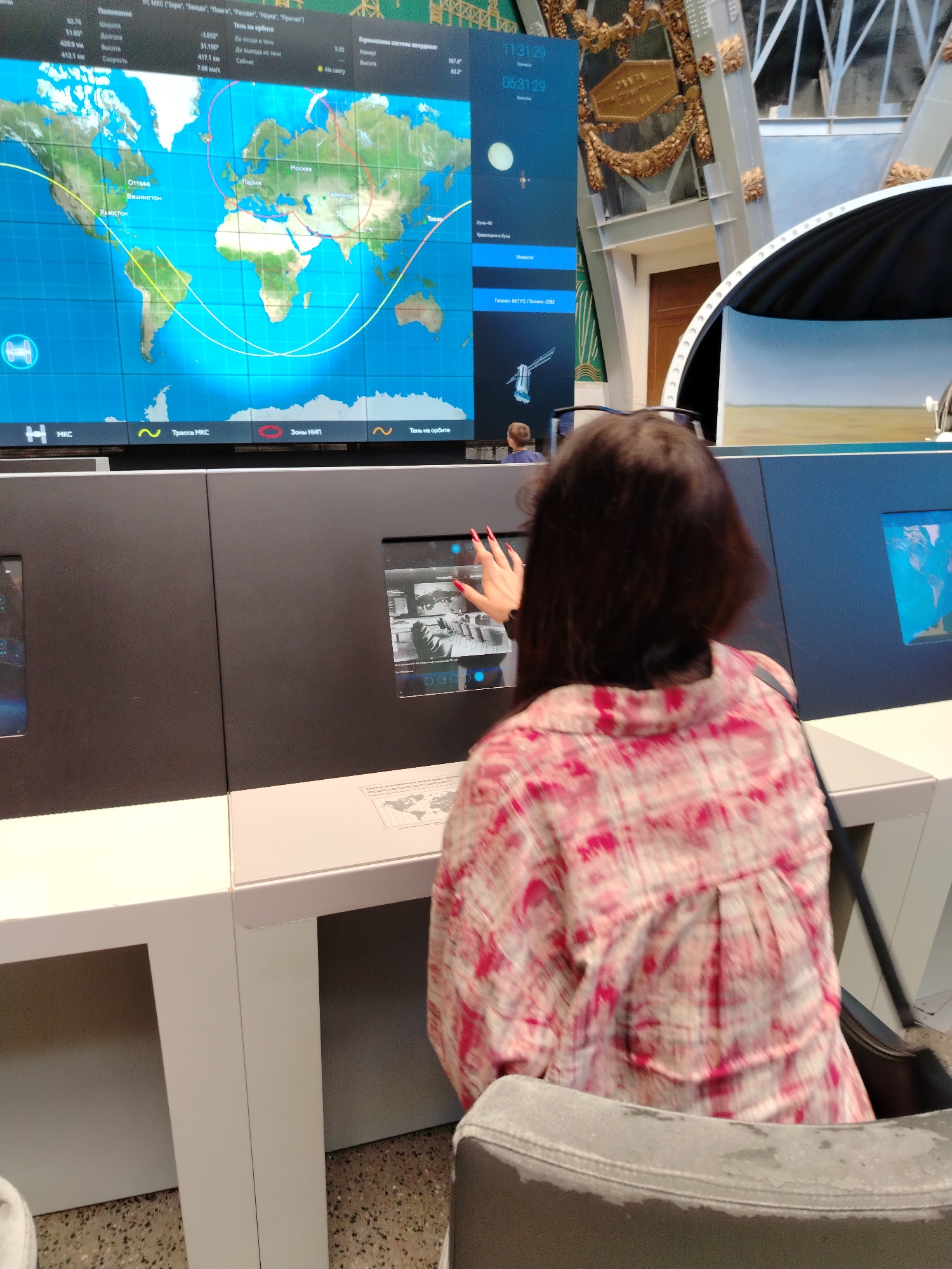 The image shows an interior view of a museum exhibit, likely related to space exploration, in Moscow, Russia. A large display screen dominates the background, showing a world map with orbital paths and geographical locations marked, along with data related to space missions. Below the large screen are several interactive touchscreens. In the foreground, a person with dark hair, wearing a pink and white patterned shirt, is interacting with one of the touchscreens. Their hand is reaching out towards the screen, suggesting they are in the process of selecting an option or viewing information. The overall environment appears to be modern and educational, with a focus on digital displays and interactive elements. The lighting is typical of an indoor exhibition space.