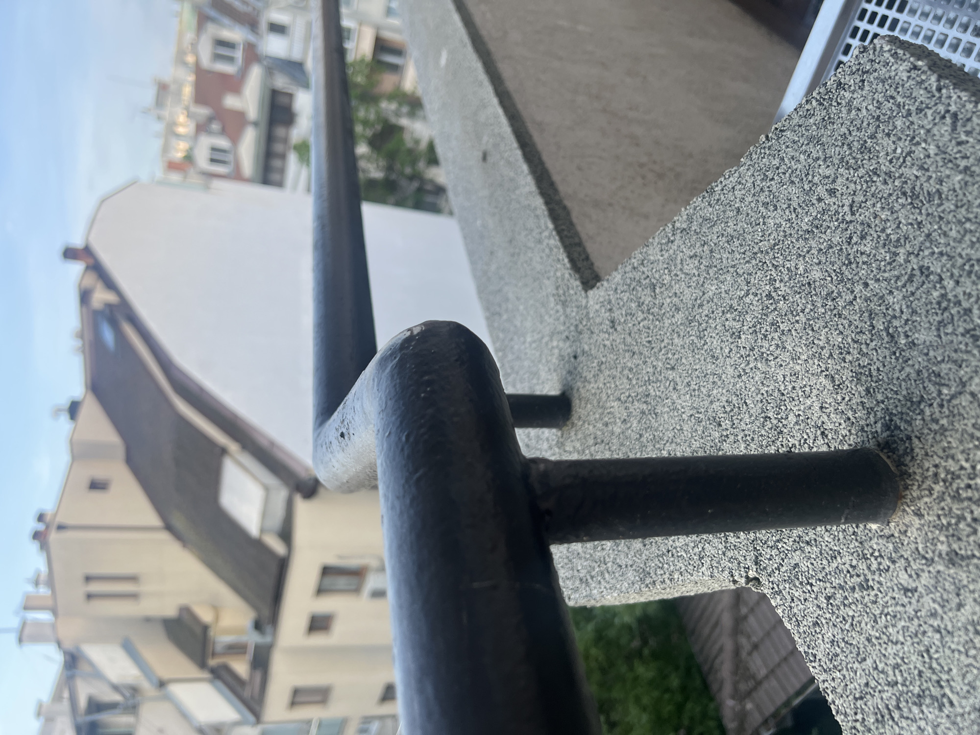 The image depicts a close-up view of a black metal railing attached to a textured, speckled grey concrete balcony. In the blurred background, buildings typical of urban architecture are visible, including rooftops with gables and various windows. The buildings are painted primarily in white and shades of beige. The sky is visible in the upper left, indicating daylight. Based on image analysis, the location is plausibly Varna, Bulgaria, based on architectural styles present. There are no people visible in the image.