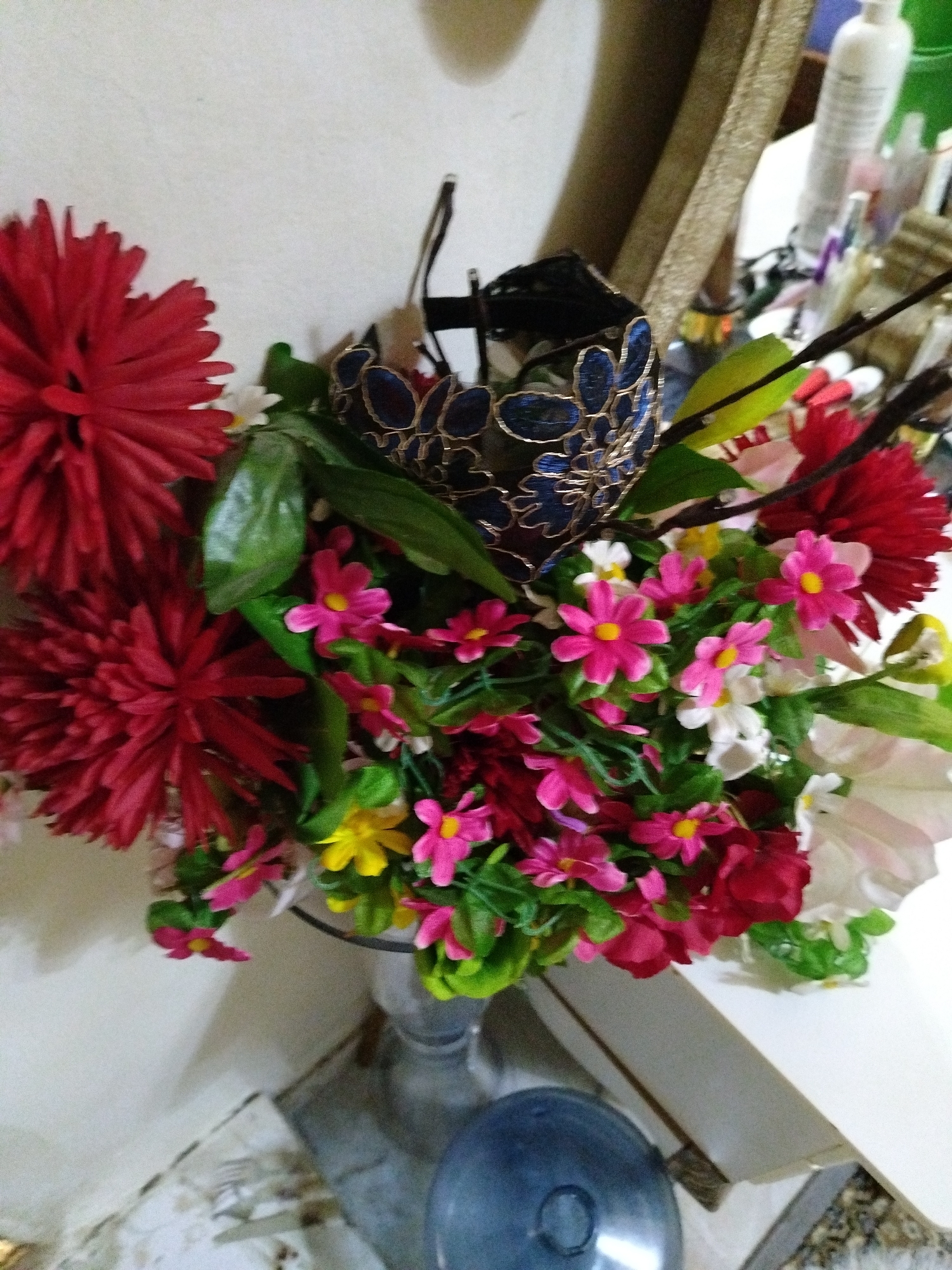 A close-up shot shows a bouquet of artificial flowers in a silver-colored vase. The flowers are predominantly red and pink, with some white and yellow blooms, and green foliage. A black and blue ornamental basket sits among the flowers. The arrangement is placed on a white surface, which may be a table or shelf. The background includes a cream-colored wall. Other items and a gold-colored object are partially visible in the background, suggesting a cluttered space. The location is not possible to determine from the information in this image.