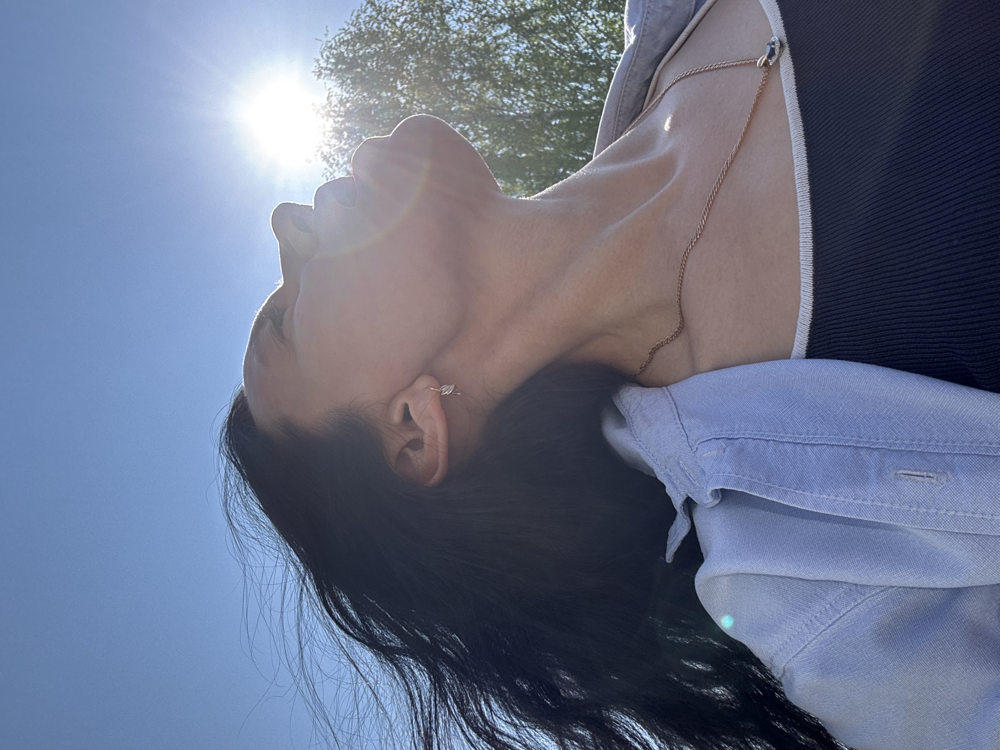 A woman with long, dark hair, an earring in her left ear, and a gold necklace is looking upwards into the sun. She is wearing a black tank top and a light blue button-down shirt. The background is a bright blue sky with a bright sun and a blurry tree in the distance. The location is Chaikovsky, Russia.