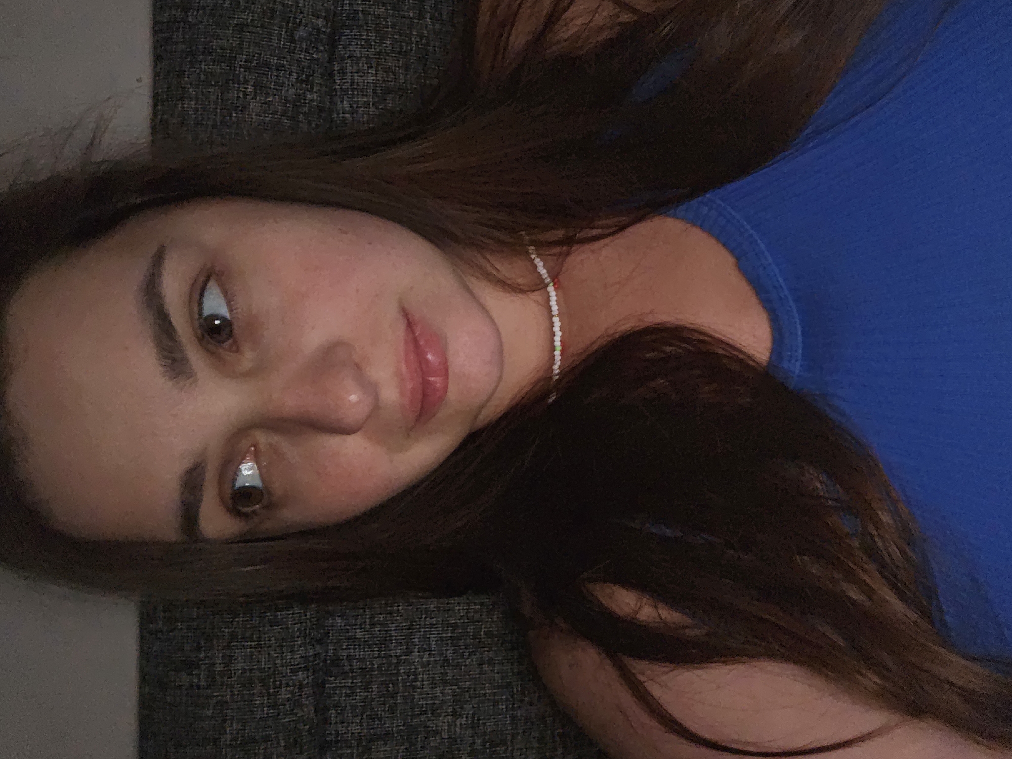 The image shows a close-up, medium shot of a young woman with long, dark brown hair, lying down and looking to her left. She is wearing a bright blue tank top and a delicate white beaded necklace. The background is a dark, textured surface, possibly a sofa or upholstery. The lighting is dim, creating a soft glow on her face. Her expression appears thoughtful or contemplative. The scene is set in a residential environment in Petropavlivska Borshchahivka, Ukraine.