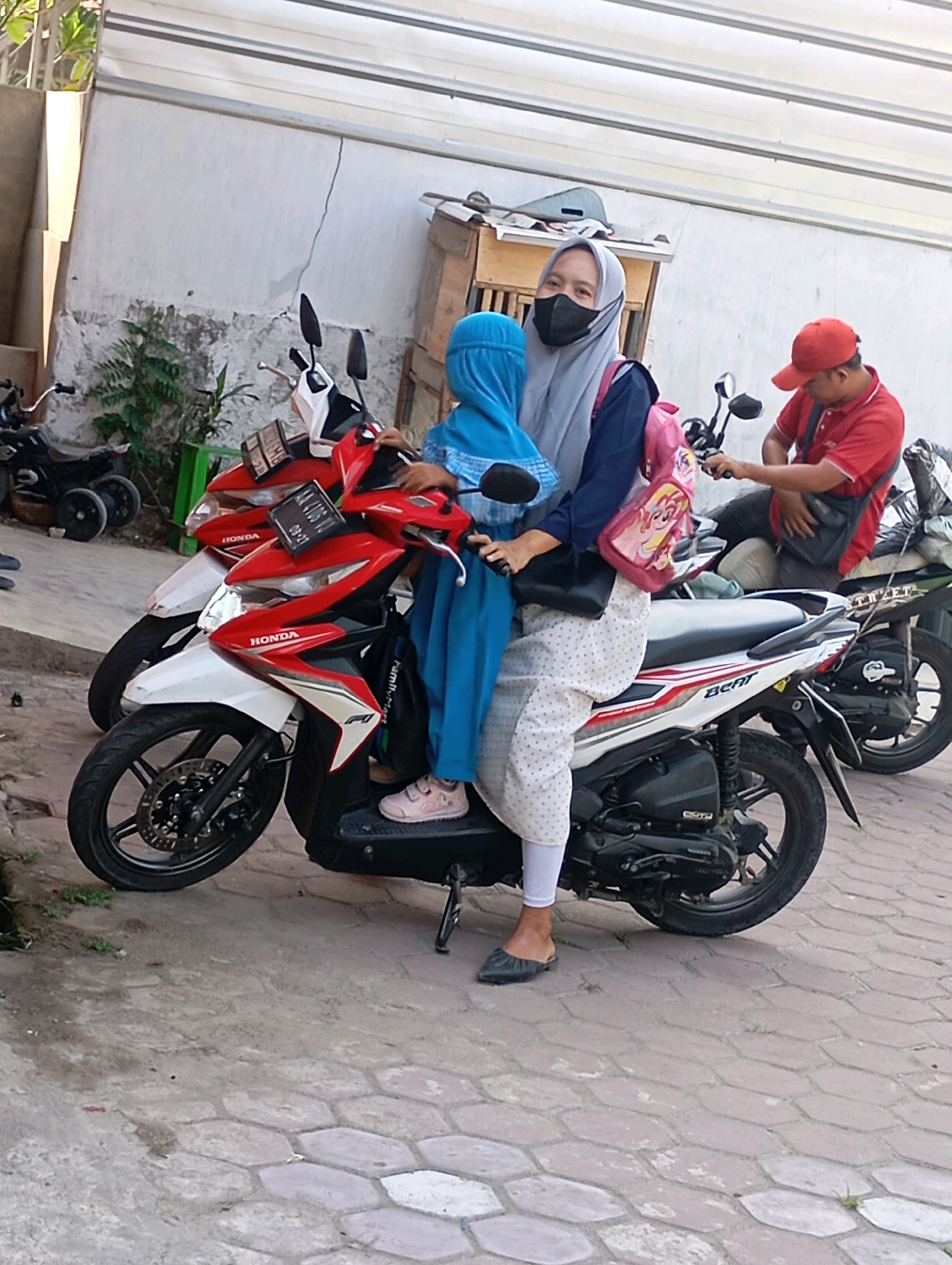 In a city setting in Babelan, Indonesia, a woman wearing a light grey hijab and black face mask is seated on a white and red Honda Beat motorcycle. She is holding a child, who is wearing a blue hijab, in front of her. The woman is wearing a dark blue long-sleeved shirt and a white polka-dot patterned skirt. She has a pink backpack with a cartoon character design on her back. Next to them, another red Honda Beat motorcycle is parked. A man wearing a red shirt and a red cap is seated on this second motorcycle. The ground is paved with hexagonal concrete blocks, and some green plants are visible in the background. The scene appears to be on a street or parking area.