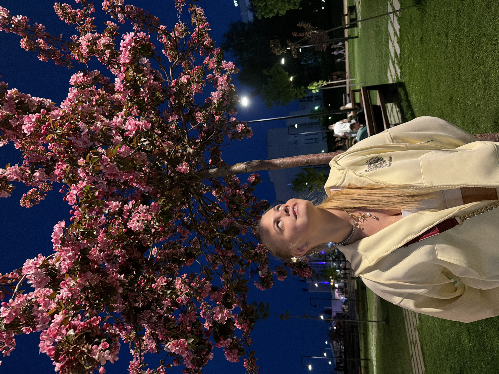 A young woman in a light yellow hoodie stands outside at night in Izhevsk, Russia, looking up at a tree with pink blossoms. The dark blue sky is visible between the tree's branches. The surrounding environment includes benches, grass, lampposts, and buildings, suggesting a park or urban area.
