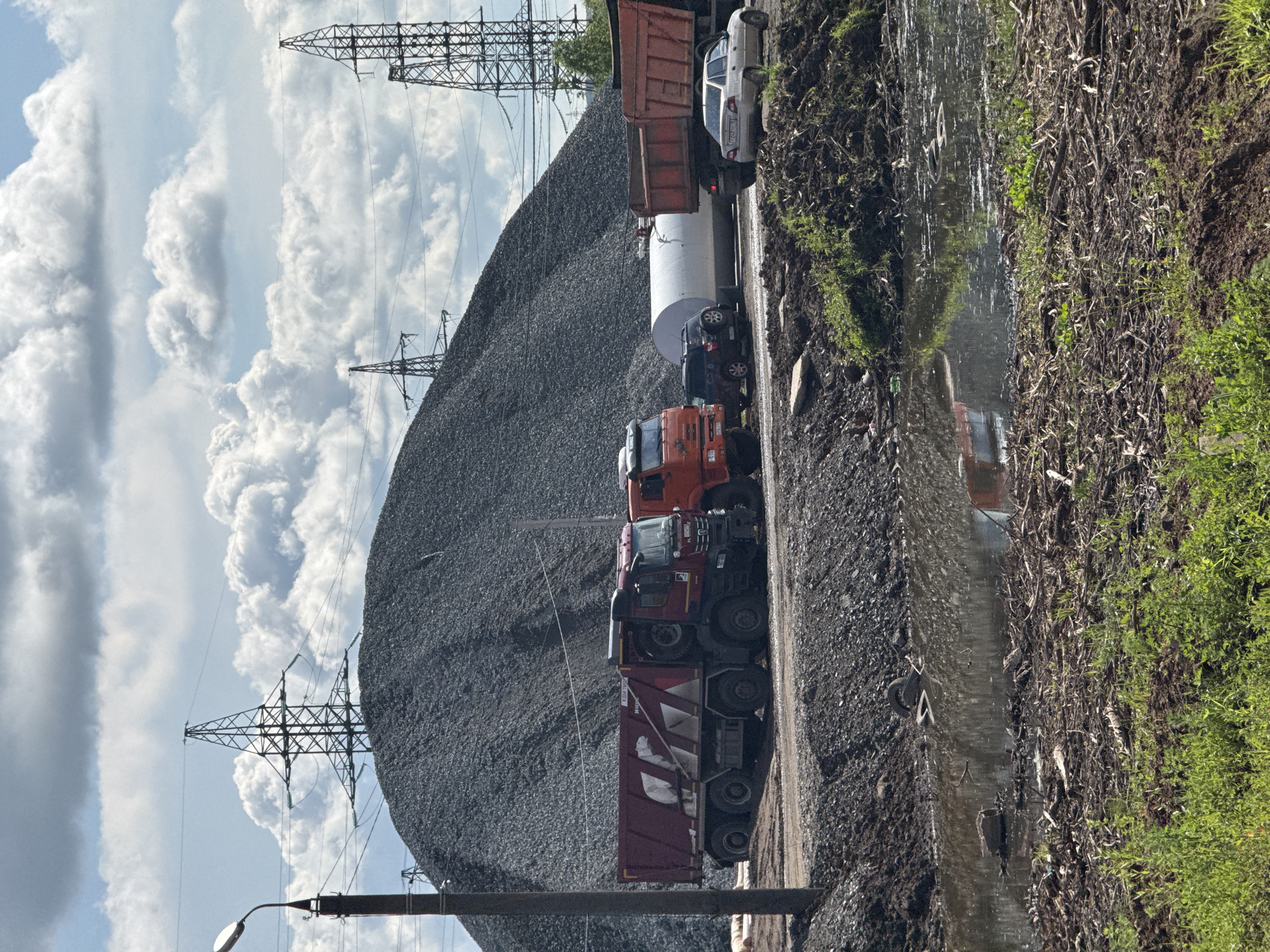 In Kabanikha, Russia, a scene unfolds at what appears to be a construction or industrial site, likely a quarry or material storage area. A massive mound of grey gravel dominates the background, with several towering electricity pylons positioned behind it, hinting at the site's proximity to power infrastructure. In the foreground, two large dump trucks are parked side-by-side: a deep red one on the left, and an orange one to its right. Further to the right, a white trailer and another dump truck are visible. A dark-colored SUV is parked behind the orange dump truck, and a light-colored sedan is parked in front of the rightmost dump truck. The ground in front of the trucks is a mixture of dirt and gravel, transitioning into a muddy, shallow body of water reflecting the sky and surrounding objects. The water is littered with debris, including branches and what appear to be discarded tires. The immediate foreground shows rough ground covered in a dense layer of twigs and low-lying green vegetation. The sky above is partly cloudy with a mix of white cumulus clouds against a light blue backdrop. There are no people visible in the scene. The overall impression is one of an active, if somewhat untidy, operational area.
