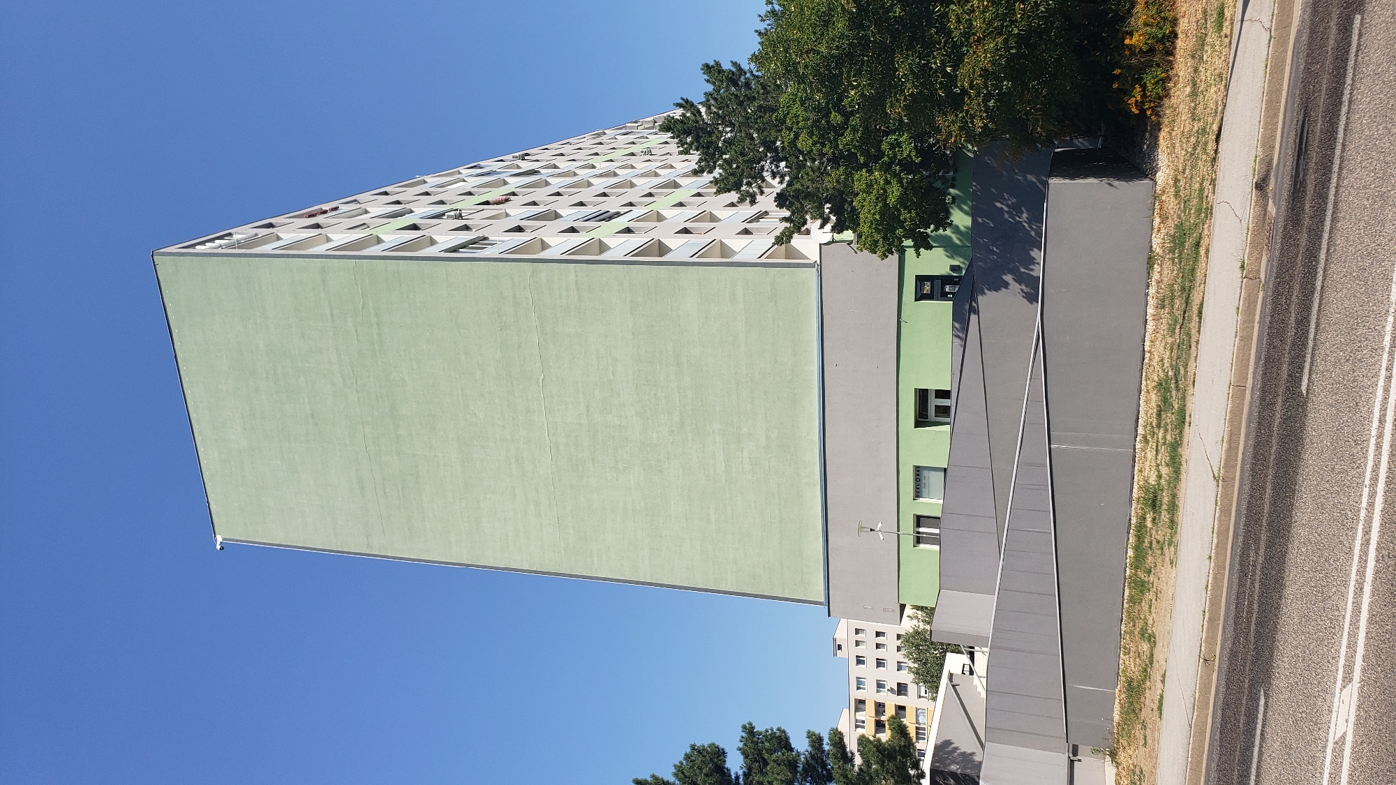An exterior, low-angle, full shot captures a tall, pale green apartment building against a clear blue sky. The building is situated in a city, likely Bratislava, Slovakia, indicated by the architecture typical of socialist-era housing. The left side of the building features a large, unadorned pale green facade, while the right side reveals a grid of windows and balconies, characteristic of a residential high-rise. A gray base surrounds the lower portion of the building, and a distinctive zigzagging gray ramp system is visible in the foreground, leading up to the building's entrance. Adjacent to the building, a dense green tree partially obscures the view of the upper floors on the right side. In the distance, another yellow and white apartment building is visible, suggesting a residential neighborhood. The street in the very front is asphalted with faded white lines, and a sidewalk with dry grass borders it. The overall scene is well-lit by sunlight, creating shadows. There are no people visible in the frame.