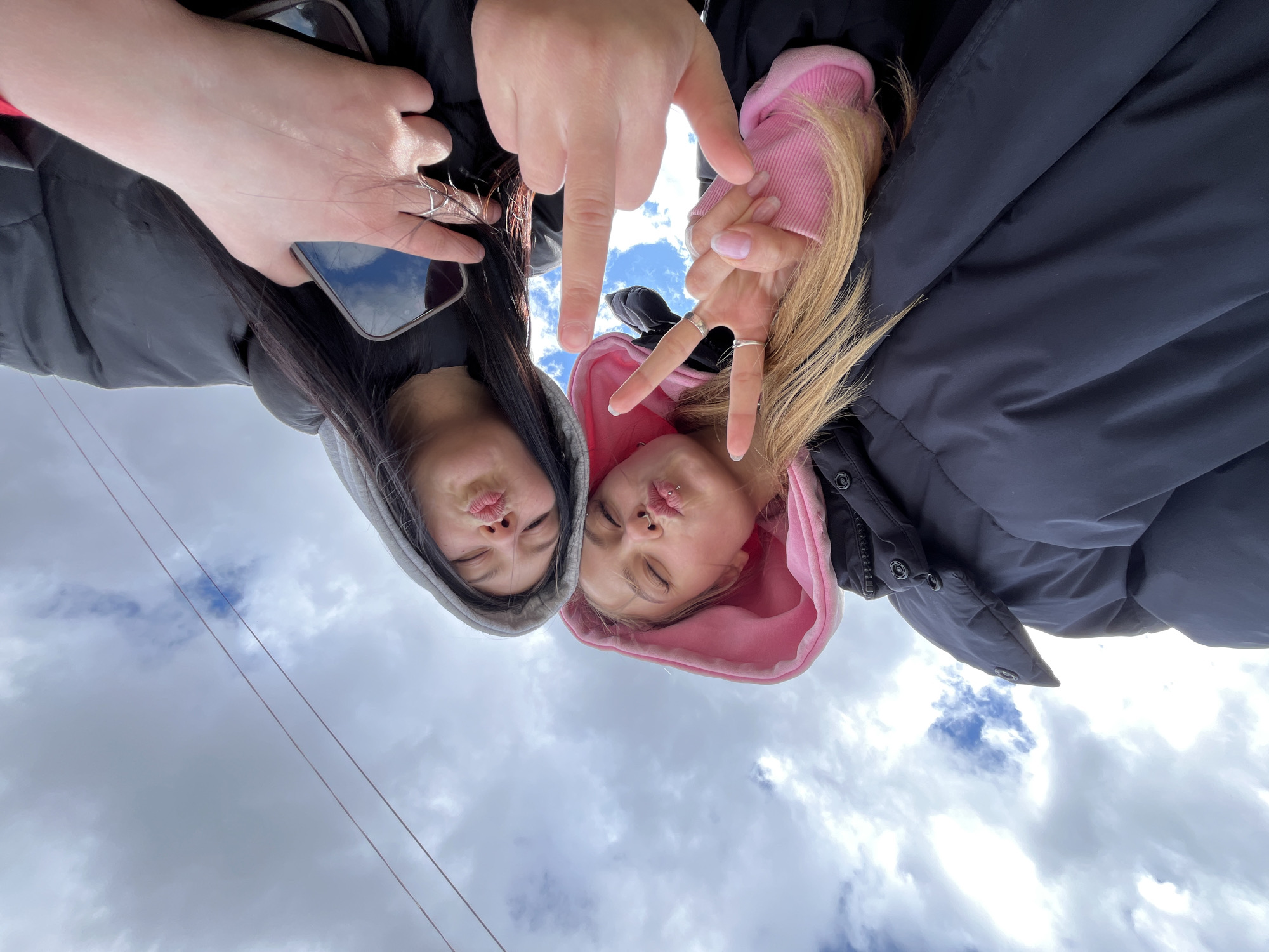 This upward-facing portrait captures two young women posing playfully against a cloudy sky in Chaikovsky, Russia. The image is taken from a low angle, looking up at the subjects, who appear to be leaning over the camera.
The woman on the left has long dark hair and wears a dark hooded top and jacket. She holds a smartphone in her left hand, which has rings on some fingers, and its screen reflects the sky. Her right hand is also raised, possibly making a peace sign, and she wears rings on that hand as well. Her eyes are closed, and her lips are pursed in a "pout" or "kissing" face.
The woman on the right has long blonde hair and wears a pink hooded sweatshirt under a dark puffer jacket. She has a septum piercing and multiple rings on her fingers. Her right hand is clearly visible making a peace sign, and her left hand is also partially visible, seemingly making a peace sign. She also has her eyes closed and lips pursed in a similar playful expression.
The background consists of a bright, overcast sky with several patches of blue visible through the clouds. Two thin, dark lines, possibly wires, run diagonally across the lower left section of the sky. The overall scene conveys a casual, friendly, and playful mood, likely captured during the daytime. There is no visible text in the image.