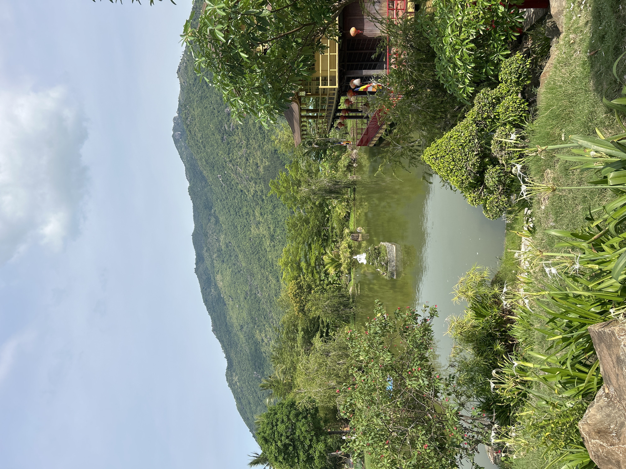 The image captures a serene view of a tranquil pond surrounded by lush greenery and a traditional building, set against a backdrop of rolling, tree-covered mountains. In the center of the pond, a small island features a white statue, possibly of a deity or a mythical figure, adorned with a small cluster of plants. The water of the pond is a muted green, reflecting the surrounding foliage and the overcast sky.
To the right of the pond, a multi-story building with a distinctive Asian architectural style stands prominently. It features a yellow exterior, a dark, sloped roof, and verandas with red railings. Lanterns are visible hanging from the eaves, adding to the cultural aesthetic. Several individuals are present on the building's balcony and near its entrance, suggesting it is a place of interest or worship.
The foreground is dominated by a variety of plants, including tall, white-flowered lilies and intricately shaped, manicured shrubs. Rocks are interspersed with the vegetation, adding a natural, earthy element to the scene. The overall impression is one of a well-maintained garden or temple complex, likely situated in a peaceful, natural environment. The location is identified as Nam Nha Trang Ward, Vietnam, known for its scenic beauty and cultural heritage.