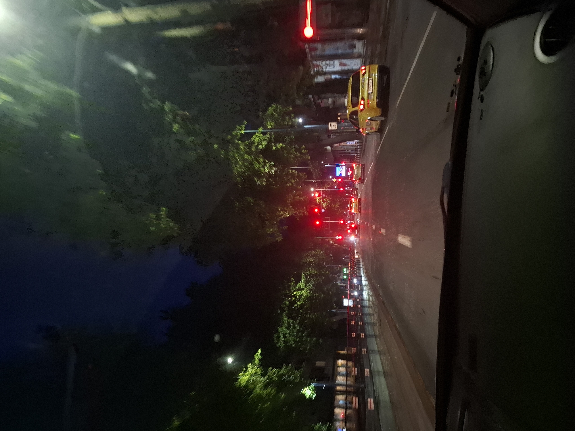 The image shows a nighttime view from inside a vehicle, looking out at a city street. The road is wet, reflecting the lights of street lamps and traffic signals. Trees line the street, their leaves illuminated by artificial lights, creating a dappled effect. Several cars are visible, including a yellow taxi in the right lane. Traffic lights are red in the distance, indicating a stop. Buildings with illuminated windows can be seen on the left side of the street. The overall scene suggests a typical urban environment at night, with ongoing traffic and ambient lighting.