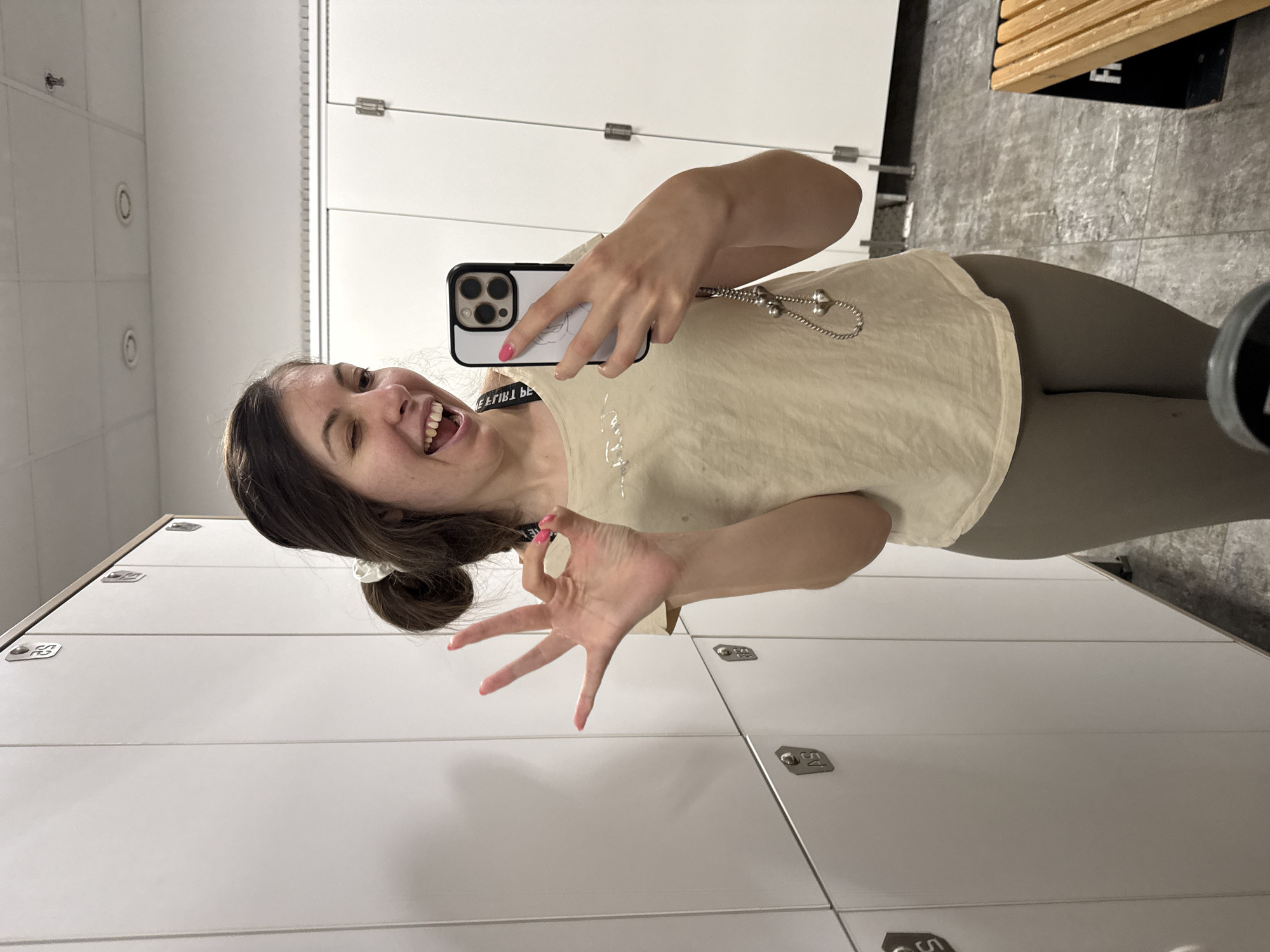 A woman with brown hair in a messy bun winks and smiles while holding up her right hand in an "okay" gesture. She is wearing a light-colored t-shirt and grey leggings. She is taking a selfie with her iPhone, which has a white case and a heart charm attached by a silver chain. The background consists of white lockers and a grey tiled floor. A wooden bench is visible to the right.