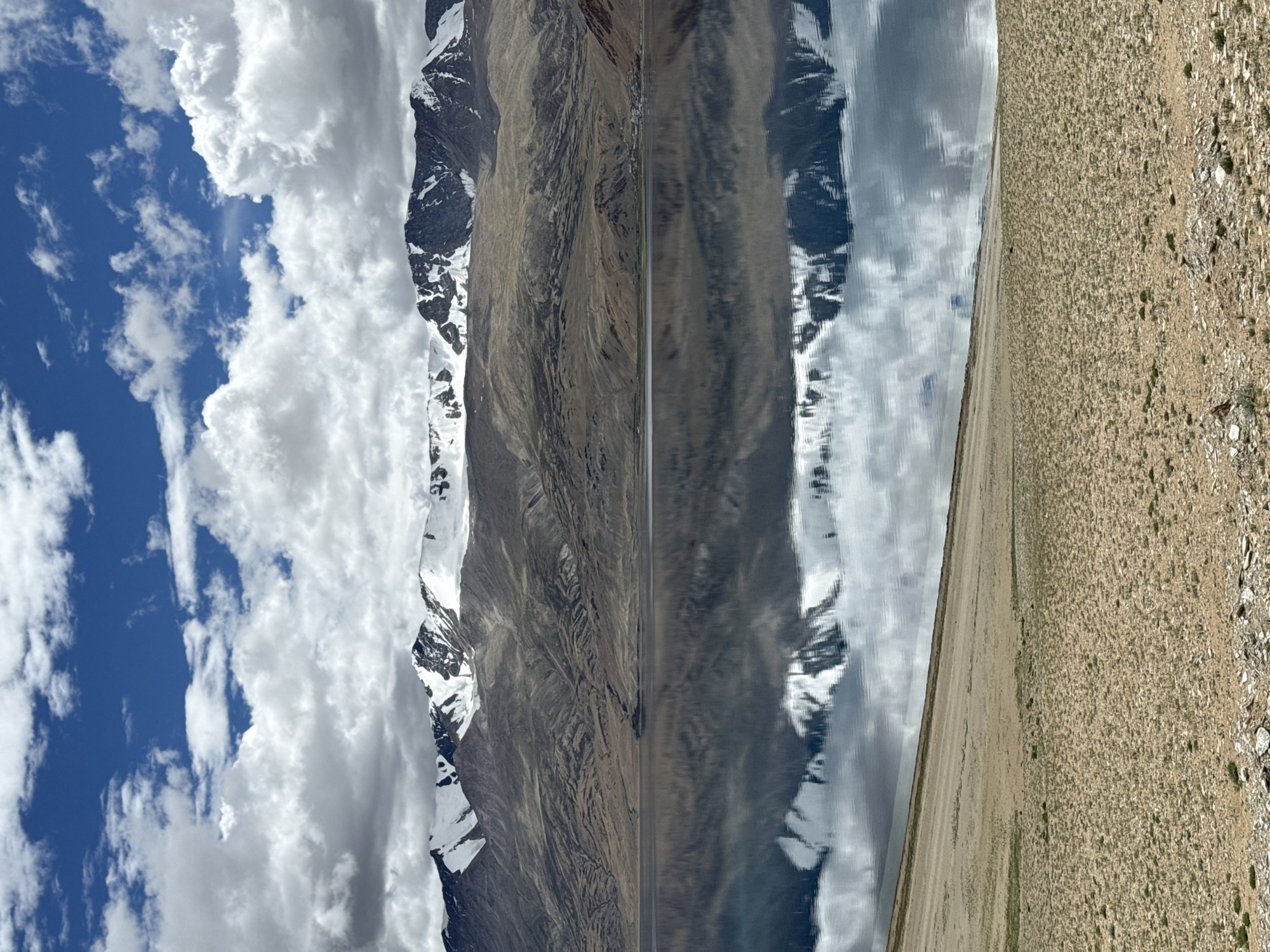 The image depicts a serene mountain landscape with a reflective lake in the foreground, located in an unknown area of India. A chain of snow-capped mountains dominates the background, their rugged peaks contrasting with the clear blue sky dotted with white clouds. The lake's surface acts as a mirror, perfectly reflecting the mountains and the sky above, creating a symmetrical and tranquil scene. The foreground consists of a gently sloping, barren terrain covered with sparse vegetation and small rocks. The lighting suggests it is daytime, possibly late morning or early afternoon, with the sun casting soft shadows. There are no people or man-made structures visible in the immediate vicinity, emphasizing the natural beauty and vastness of the environment.
