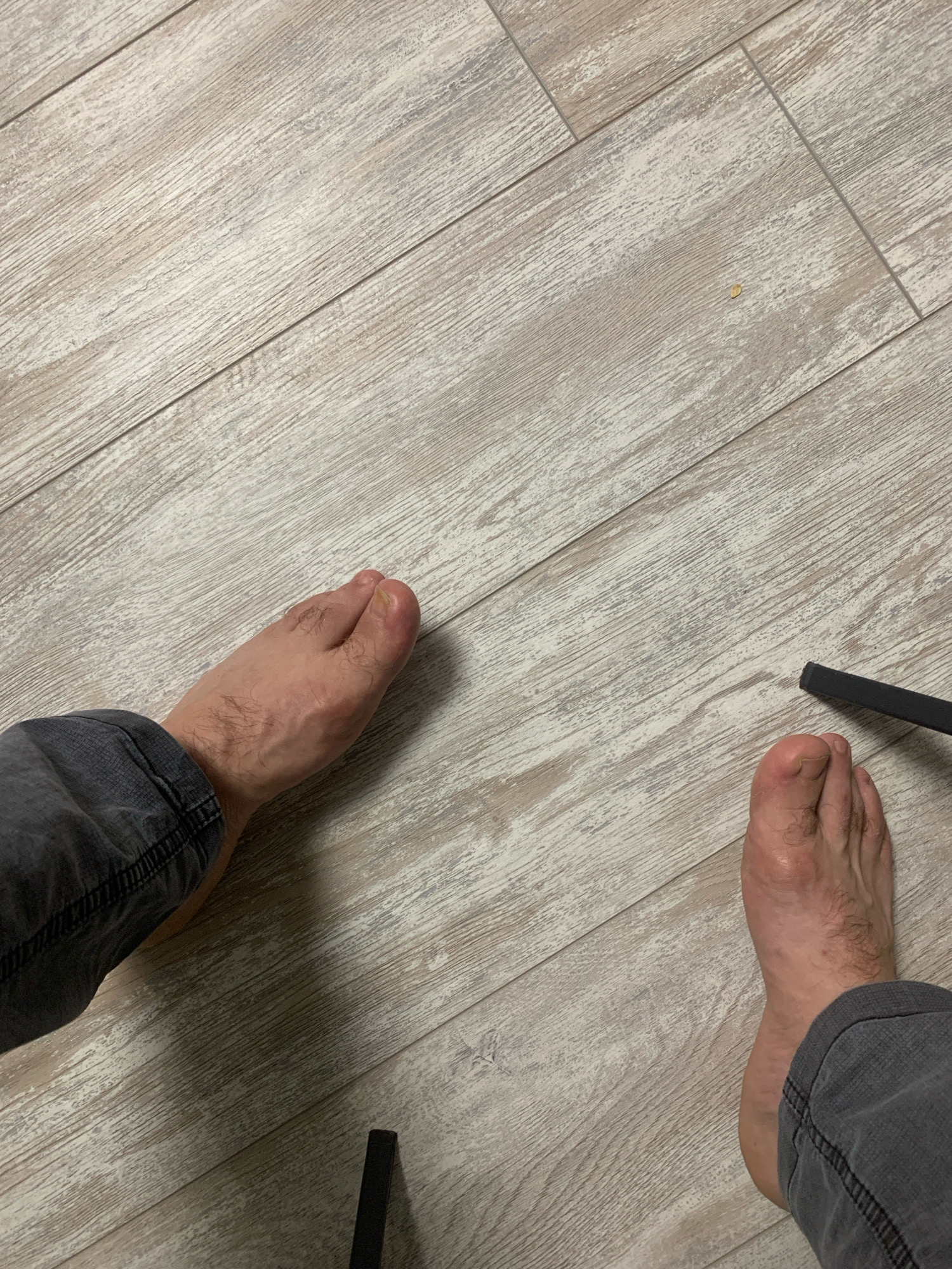 The image displays a close-up, overhead view of two bare feet positioned on a light-colored wooden floor. The feet are of a light-skinned individual, and the left leg, clad in dark gray trousers, is visible on the left side of the frame. The right leg, also in dark gray trousers, is visible on the right side of the frame. The floorboards are laid diagonally across the image, exhibiting a distressed wood grain pattern with variations in light and dark tones. A dark, slender object, possibly part of a piece of furniture or a tool, is partially visible in the lower right corner of the image. The scene is static and does not depict any discernible action or interaction. The environment appears to be indoors, likely a residential or commercial space with a clean, well-maintained floor. There is no information in the image to determine its location in the city of Kyakhulay, Russia.