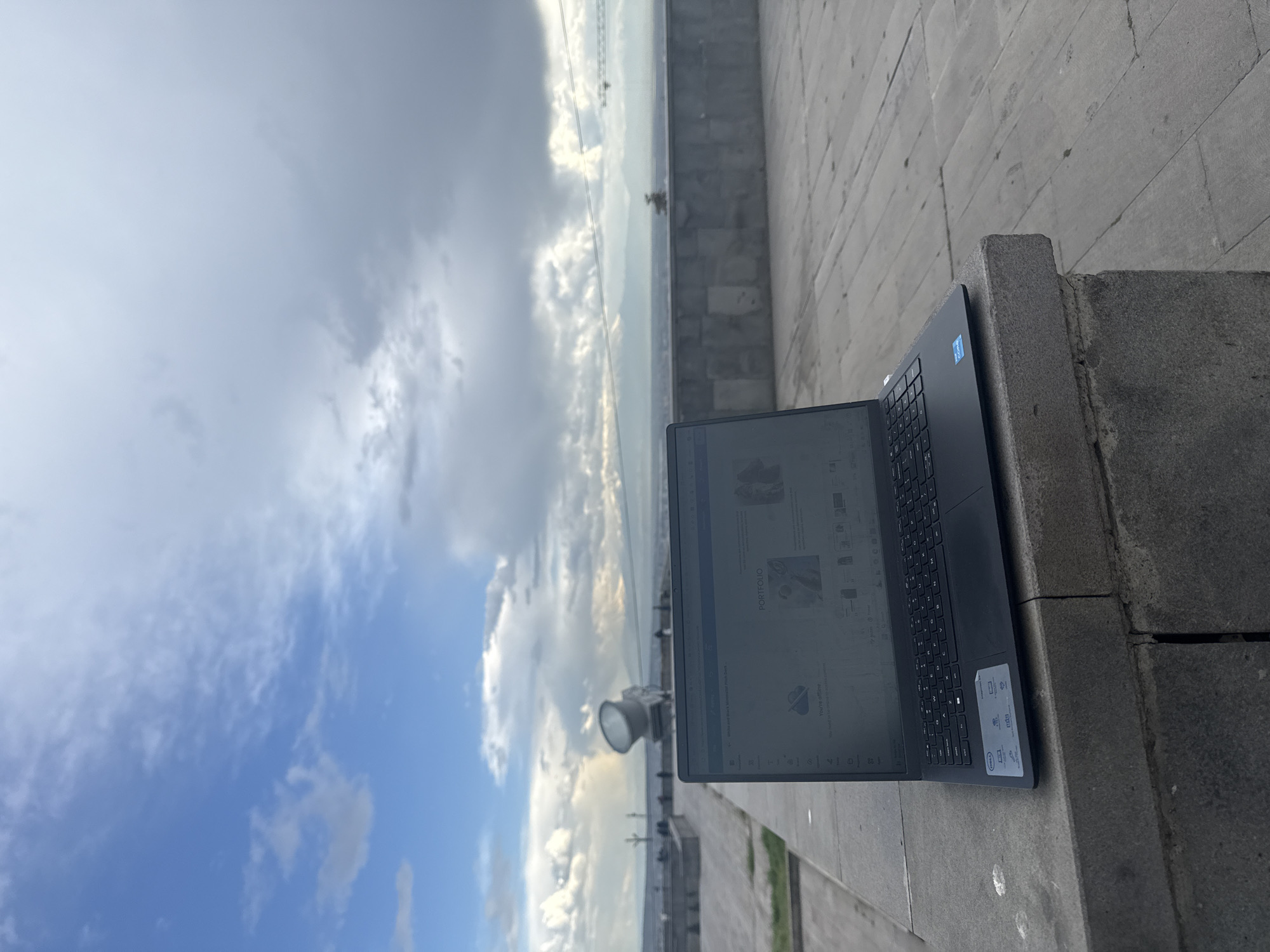 A laptop is sitting on a concrete ledge overlooking a cityscape with a cloudy sky. The screen of the laptop displays a website with the word "PORTFOLIO" visible. The cityscape includes tall structures resembling bridges or towers in the distance. The sky is a mix of blue and grey clouds. The location is Yerevan, Armenia.
