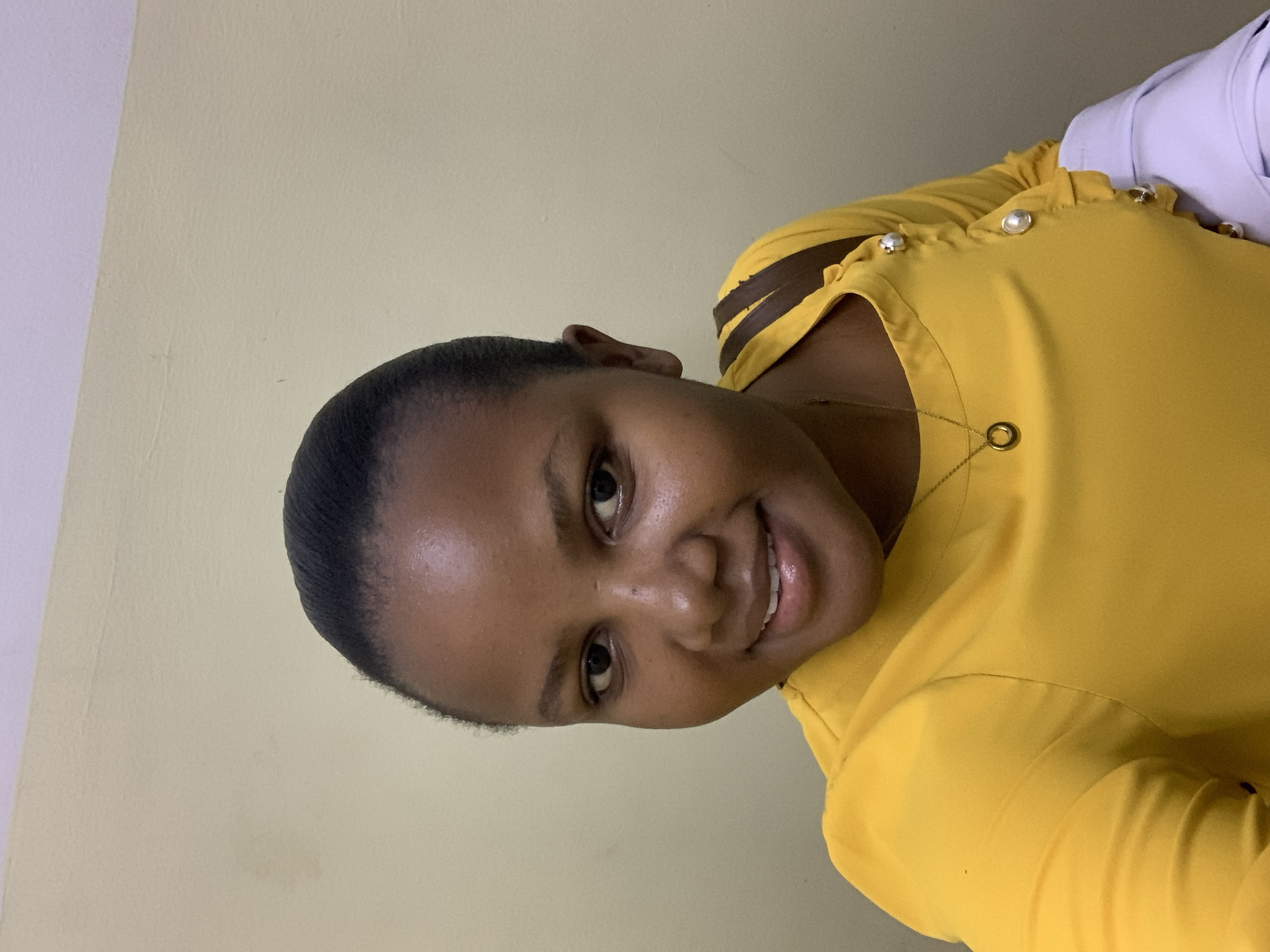 A young woman with dark skin is shown from the shoulders up, turned slightly to the left, smiling, and looking directly at the viewer. She is wearing a yellow blouse with decorative pearl-like buttons on the upper right shoulder. A gold necklace with a circular pendant is visible. Her hair is short and dark. The background is a light yellow wall. The location is unknown and cannot be determined from the image.