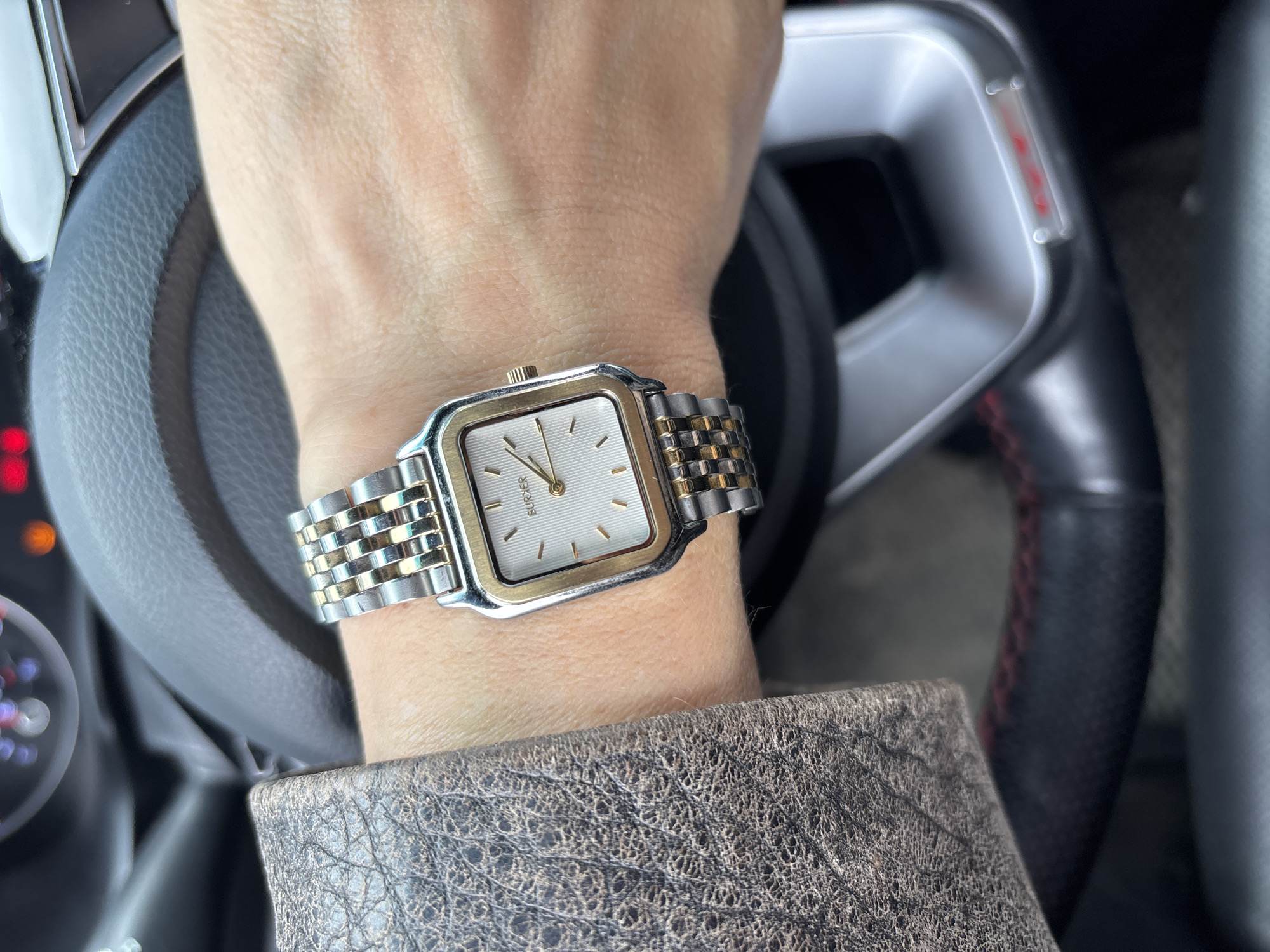 A person's wrist is displayed wearing a two-tone metal watch with a square face. The hand is resting on a car steering wheel. The person is wearing a patterned jacket.