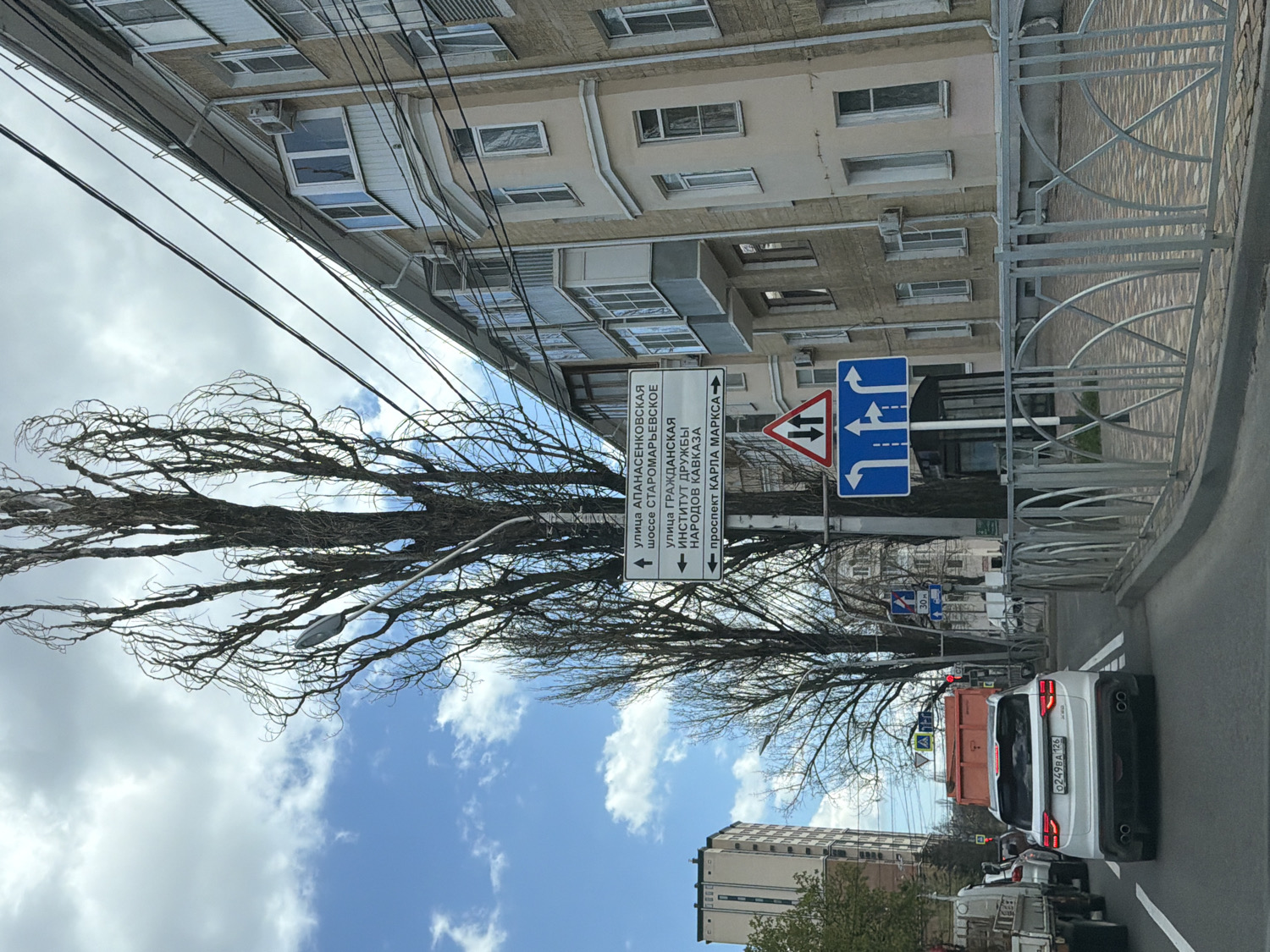 The image captures a daytime urban street scene, likely from a vehicle, in a Russian city. The sky is partly cloudy with blue patches visible between dark, tangled branches of a large, bare-limbed tree on the left. Utility wires traverse the upper part of the frame.
On the right, a multi-story, light-colored apartment building with numerous windows and balconies stands behind a decorative metal fence. Several road signs are prominent near the building. A white rectangular sign with Cyrillic text and arrows provides directions to various locations: "улица АПАНАСЕНКОВСКАЯ" (Apanasenkovskaya street), "шоссе СТАРОМАРЬЕВСКОЕ" (Staromaryevskoe highway), "улица ГРАЖДАНСКАЯ" (Grazhdanskaya street), "ИНСТИТУТ ДРУЖБЫ НАРОДОВ КАВКАЗА" (Institute of Friendship of Peoples of the Caucasus), and "проспект КАРЛА МАРКСА" (Karl Marx Avenue). Adjacent to it are blue mandatory traffic signs indicating lane usage: one shows an arrow for straight and left turns, and another shows options for straight, left, and right turns from multiple lanes. A red-bordered triangular sign warns of two-way traffic.
In the foreground, the rear of a white SUV is visible, bearing the Russian license plate "0249BA 126". Behind the SUV, part of an orange truck can be seen. The bare tree suggests the season is either late autumn, winter, or early spring. The visible Cyrillic text and the license plate's region code (126 for Stavropol Krai) indicate the location is in Stavropol Krai, Russia.