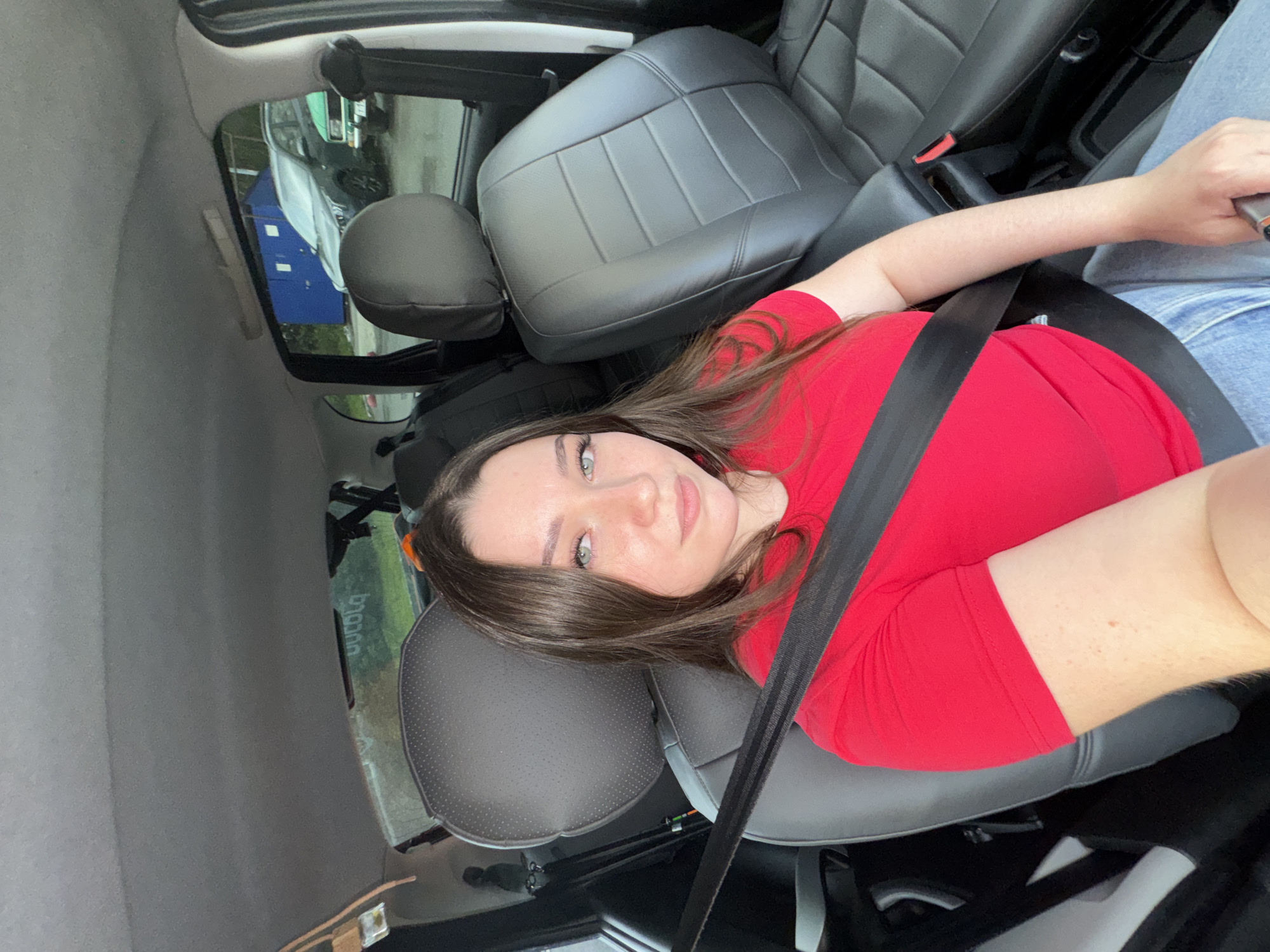A young woman is sitting in the driver's seat of a car, looking at the camera. She has long brown hair and blue eyes, and is wearing a red t-shirt and blue jeans. The car is a dark color and appears to be parked in an urban setting. In the background, a blue building is visible, as well as other parked cars. The interior of the car has black leather seats and a black seatbelt. A sticker with the word "unicola" is visible on the rear windshield. The image appears to have been taken during the daytime.