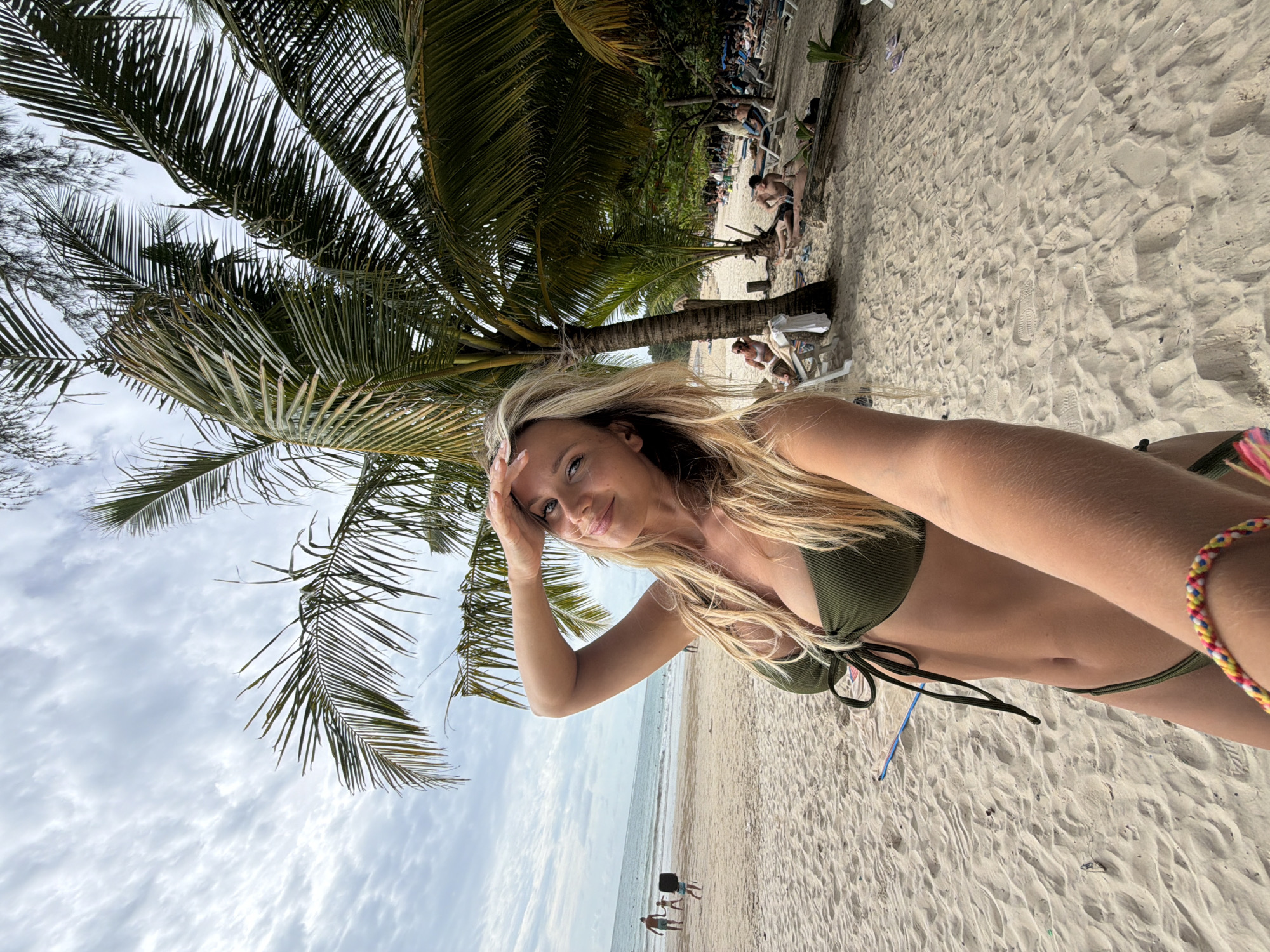 In Sakhu, Thailand, a blonde woman in a green bikini is taking a selfie on a sandy beach. She is holding her hand to her forehead as if shielding her eyes from the sun. Behind her, palm trees cast shadows on the sand. The ocean is visible in the background, with gentle waves rolling onto the shore. Several people are scattered on the beach, some lounging on chairs and others walking near the water's edge. The sky is partly cloudy, with streaks of white clouds against a pale blue backdrop. The overall scene suggests a relaxed day at a tropical beach.
