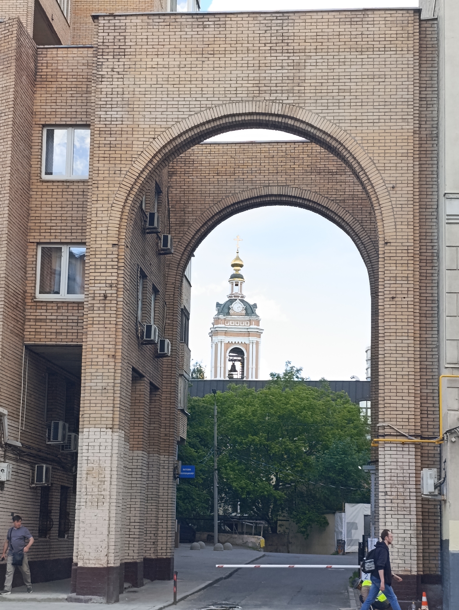 This media file captures an urban scene in Moscow, Russia, framed by a large, light brown brick archway connecting two sections of a building.
Through the arch, a traditional Orthodox church bell tower is visible in the middle ground, topped with a golden dome and a cross. The tower features a clock face and has a reddish-brown and white facade. Below the tower, green trees partially obscure other structures. The sky above is bright and overcast, indicating daytime.
To the left of the arch, a multi-story building section is visible, also constructed of light brown brick, featuring white-framed windows and several outdoor air conditioning units mounted on its exterior. To the right of the arch, another part of a building with similar brickwork and a gray panel is seen, with a yellow gas pipe running vertically.
In the foreground, two individuals are present. On the left, a man wearing a gray shirt and light trousers with a dark shoulder bag walks away from the viewer on a paved area beneath the building. On the right, another man, dressed in a dark hoodie, blue jeans, and carrying a backpack, walks across an asphalt road. He is passing a white road barrier marked with the number "2" and is carrying a yellow vest. Concrete bollards and a red traffic cone are also visible on the roadside.
A blue street sign is mounted on the wall to the left of the arch's opening, bearing the Cyrillic text "НАГАТИНСКАЯ" (Nagatinskaya), which refers to a district or street in Moscow. The overall impression is a typical urban setting on a clear day.
