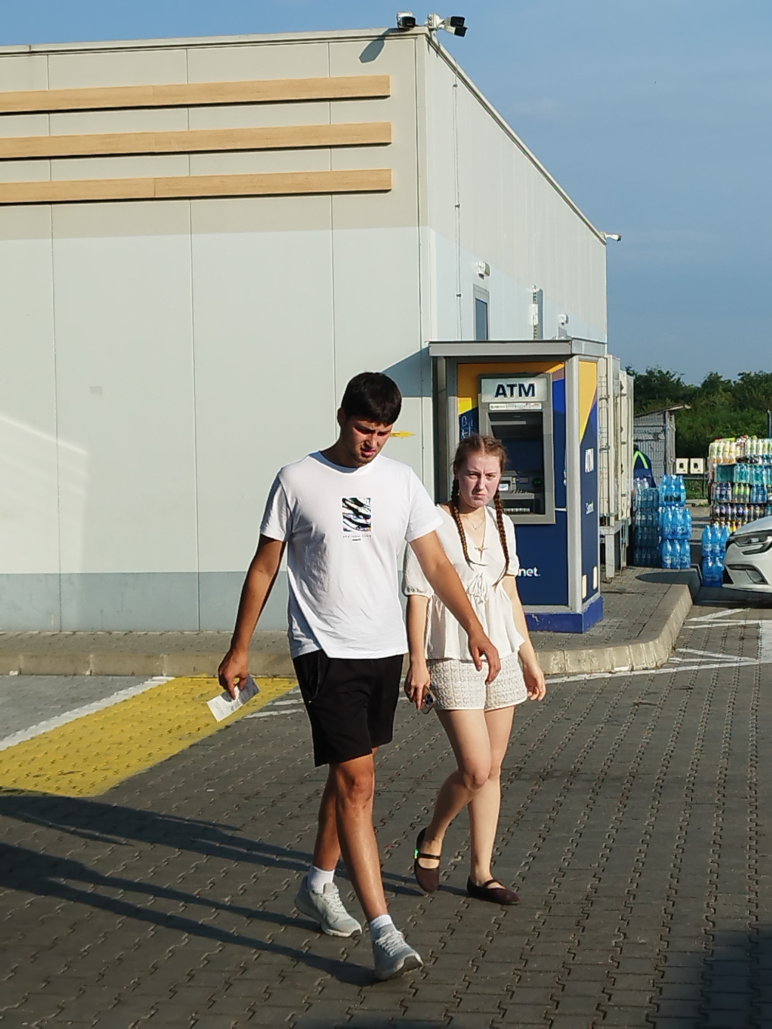 Two individuals, a male and a female, are walking on a paved area next to a modern building with a light-colored facade and horizontal wooden accents. The male, on the left, is wearing a white t-shirt and black shorts, carrying a piece of paper in his left hand. The female, on the right, is wearing a white top and textured shorts, with her hair in braids. Behind them, an ATM kiosk is visible, and further in the background, there are stacks of bottled water and a white vehicle. The scene appears to be outdoors, possibly in a commercial or service area. The specific location in Cupele, Romania, cannot be determined from the visual information provided in the image.