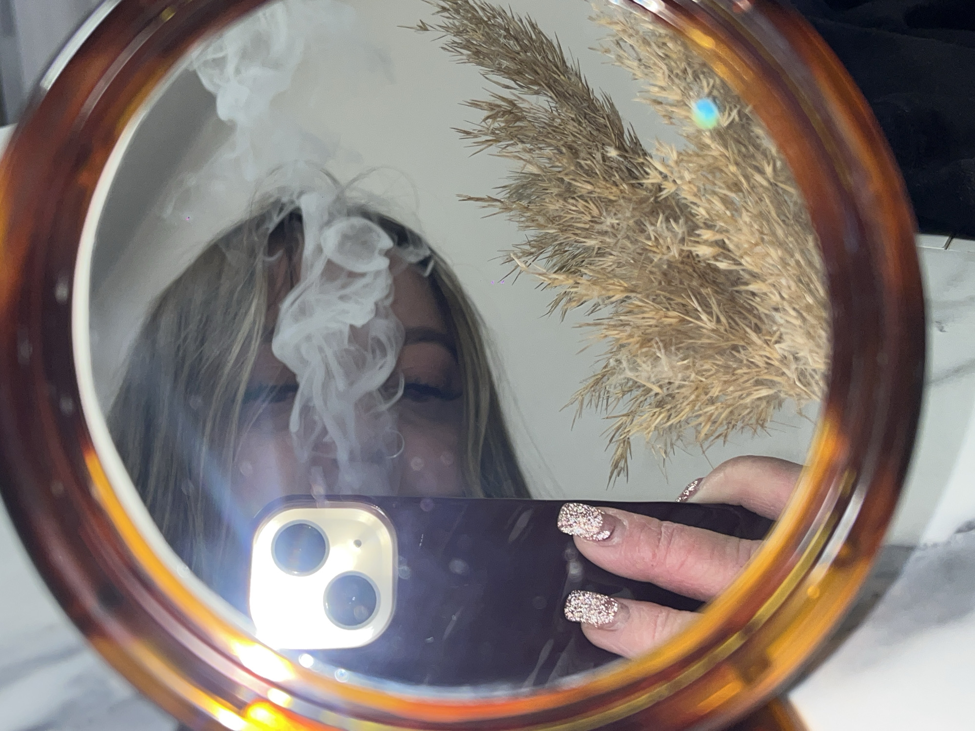 The image displays a mirror selfie, capturing a person's reflection within a round mirror with a tortoiseshell-patterned frame, set indoors in Krasnodar, Russia.
The primary subject in the reflection is a person, likely female, whose face is partially obscured by a rising plume of white smoke or vapor. Their eyes are mostly closed, and light-colored hair frames their face. They are holding a dark-colored smartphone, identifiable by its triple-camera setup, which emits a bright light, possibly a flash or screen glare. The hand holding the phone features manicured nails with sparkly, light-colored polish.
To the right within the mirror's reflection, a cluster of dried, fluffy ornamental plants, resembling pampas grass, is visible. The background further shows indistinct light-colored surfaces and a dark object, suggesting an indoor setting like a room or bathroom. Bright lens flares and reflections from light sources are scattered across the image, particularly on the phone and the mirror's surface. The activity depicted appears to be a casual selfie while exhaling smoke or vapor. No visible text is present in the image.
