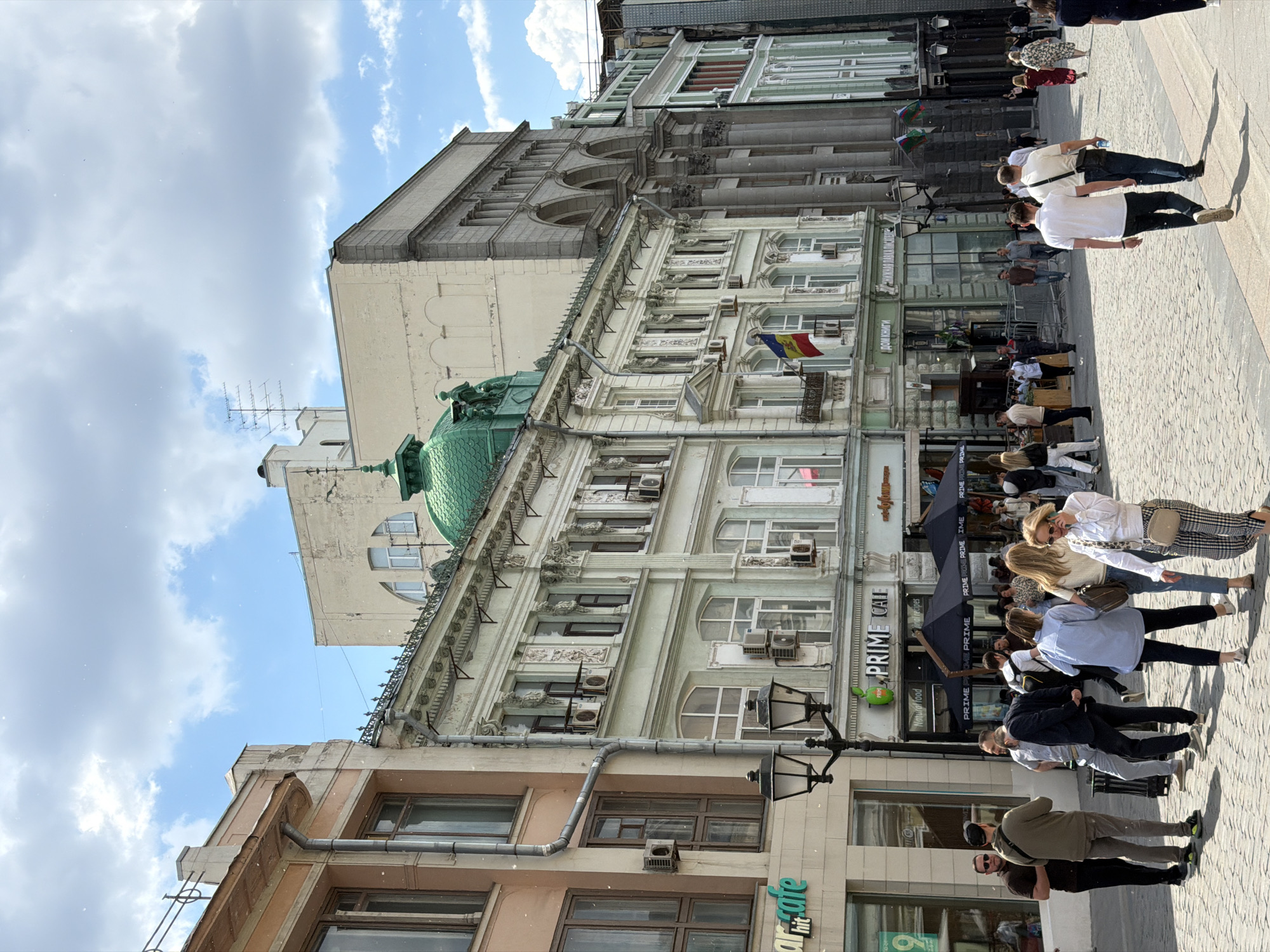 This image depicts a bustling street scene in Moscow, Russia, captured on a bright, partly cloudy day. Numerous pedestrians are seen walking and gathering along a paved street lined with ornate, multi-story buildings. The architecture features classical elements, intricate details, and a mix of light-colored facades. One prominent building has a distinctive green dome, while others show exposed brickwork and multiple windows, many equipped with air conditioning units.
People are engaged in various casual activities; some are strolling in groups or individually, while others are seated at outdoor cafe tables under black umbrellas. The clear shadows suggest it is midday or early afternoon. The atmosphere appears relaxed and lively.
Notable details include a Moldovan flag hanging from a balcony on one of the buildings, and another flag, possibly Spanish, seen further down the street. Antennas are visible on a rooftop in the background.
Several pieces of text are legible on storefronts and cafe signage. These include "PRIME CAFE", "Natural food", "hit cafe" (partially visible), and the word "PRIME" repeated multiple times on the umbrellas. Most notably, the Cyrillic text "дом книги" (Dom Knigi), meaning "House of Books," is clearly visible on one of the buildings, confirming the urban setting and location context.