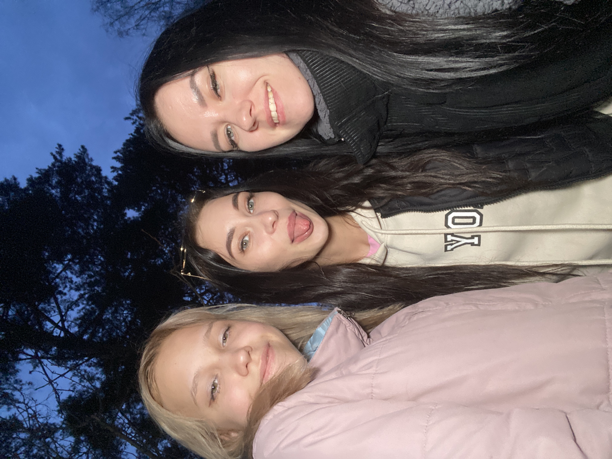 This media file features three young women posing for a low-angle selfie outdoors in Chaikovsky, Russia. The scene is set during dusk or night, as indicated by the dark blue sky and the silhouettes of tall trees in the background, suggesting a forest or park.
The women are positioned vertically, looking down towards the camera. The woman at the top has long, dark hair and is smiling, wearing a dark, possibly padded, jacket. The middle woman, also with dark hair, is playfully sticking out her tongue; she wears a light-colored hooded sweatshirt with "YO" and a blocky letter or logo (possibly a "G" inside a square) visible on it. The lowermost woman has blonde hair and a subtle smile, dressed in a light pink puffer jacket. All three appear to be in good spirits, capturing a moment together.